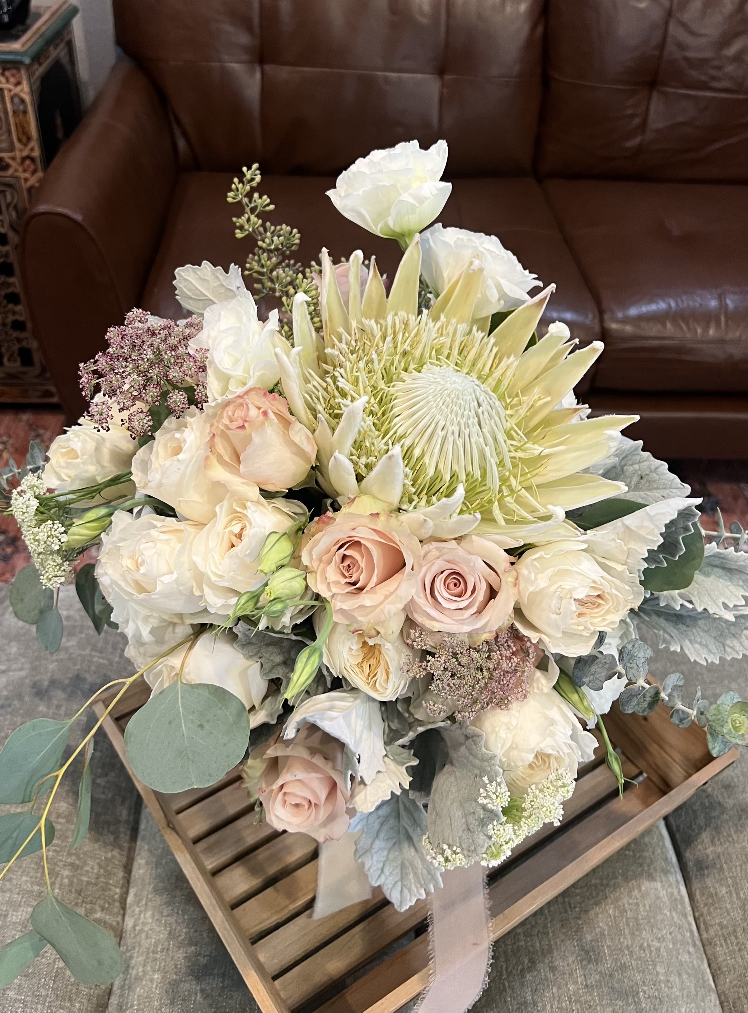 Peachy Pastel  - A pale protea-like focal bloom anchors this Peachy Pastel flower arrangement, surrounded by blush and ivory roses that create a soft, romantic statement perfect for a wedding bouquet, anniversary gift, or a formal sympathy arrangement. The gentle wash of pale pink, creamy white and hints of soft yellow reads refined and calming, the kind of bouquet that makes the person you're thinking of feel truly seen and comforted.  Delicate airy white filler flowers and soft eucalyptus with silvery foliage give the composition a garden-style texture and a muted, sophisticated silhouette. Hand-tied and finished with a ribbon wrap, the bouquet feels intimate and effortless-roses bring classic romance while the protea-like bloom adds an unexpected, elegant focal point. Ideal for brides, partners marking a milestone, or anyone needing a graceful expression of sympathy; order sizing and care details are available at checkout so the bouquet arrives fresh and ready to impress.