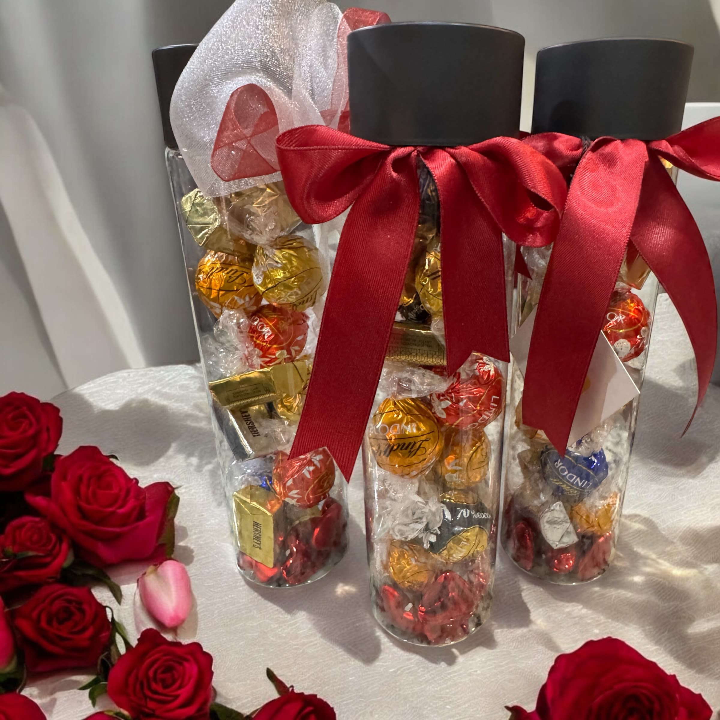 Clear gift tubes filled with chocolates and tied with red ribbons beside red roses
