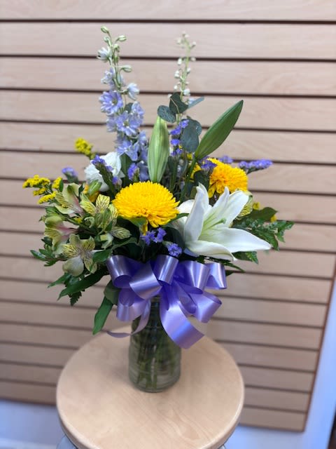 Marley - Sunlit yellow chrysanthemums and pure white Asiatic lilies create a cheerful, upright bouquet that feels both fresh and a little refined - perfect as a birthday bouquet, a spring surprise, or a bright "welcome baby" gift. The design's taller delphinium spires and lavender-toned accents lift the arrangement so the recipient will feel genuinely celebrated the moment they see it.  This mixed-color arrangement pairs alstroemeria and other seasonal blooms with airy purple-blue accents in a clear glass vase, finished with a wide purple satin ribbon for a polished touch. The composition is upright and airy with a fuller center and taller elements above; sizing fits standard vase arrangements for tabletops and entryways. Choose this when you want a thoughtful thank-you, a spontaneous "just because," or a sunny seasonal pick-me-up that keeps looking graceful for days.