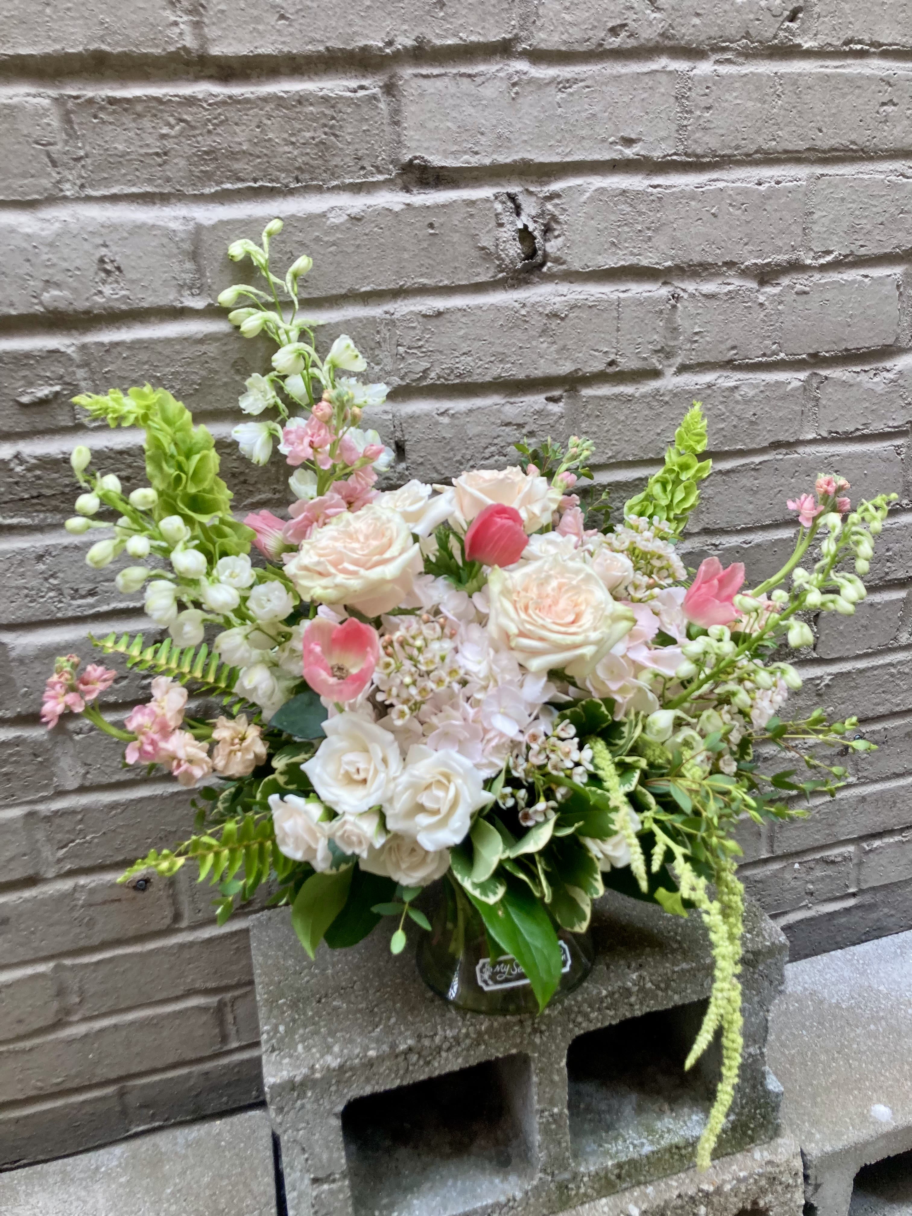 Deja Rosè - Soft blush roses and airy garden blooms create a delicate, romantic statement-perfect as an anniversary arrangement, wedding-inspired centerpiece, or a thoughtful gift. This flower arrangement for birthdays or summer celebrations carries a gentle palette of white, pink, peach and fresh green that makes the recipient feel cherished and truly seen.  Built in a clear glass vase, Deja Rosè is a large, loose garden-style arrangement with cream and blush roses at the heart, complemented by, hydrangea accents, delphinium, stock and dainty fillers for height and texture. The design blends dense, rounded clusters at the base with tall branching stems that extend outward, giving a hand-arranged, natural silhouette and abundant foliage for contrast.  Ideal for someone who appreciates an elegant yet relaxed floral look-whether you're saying "happy birthday," "congratulations," or "I'm thinking of you"-this lush mixed bouquet arrives ready to display in the included glass vase. For best enjoyment, place in a cool spot away from direct sun and refresh the water every two to three days to keep blooms looking fresh.