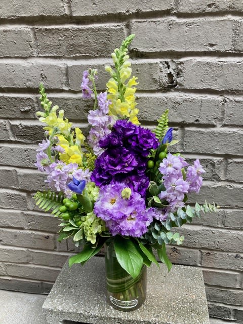 Lovely Lisianthus - Tall lavender and purple blooms mingle with sunny yellow accents in this garden-style flower arrangement, perfect as a cheerful birthday bouquet, spring celebration centerpiece, or just-because thank-you gift. Upright snapdragon spikes create graceful height while rounded clusters of hydrangea and locally grown Lisianthus blossoms form a lush, textured center that will make the recipient feel noticed and brightened.  Carefully hand-arranged in a clear glass vase, the mix includes snapdragons, lisianthus, stock and supporting green filler for a layered, airy silhouette. The palette of purple, lavender, yellow and green reads fresh and lively; the visible stems in the glass vase add a simple, slightly rustic charm. Sized to sit on a table or desk, this composition arrives ready to display-just add fresh water and trim stems a little every few days to extend its life.