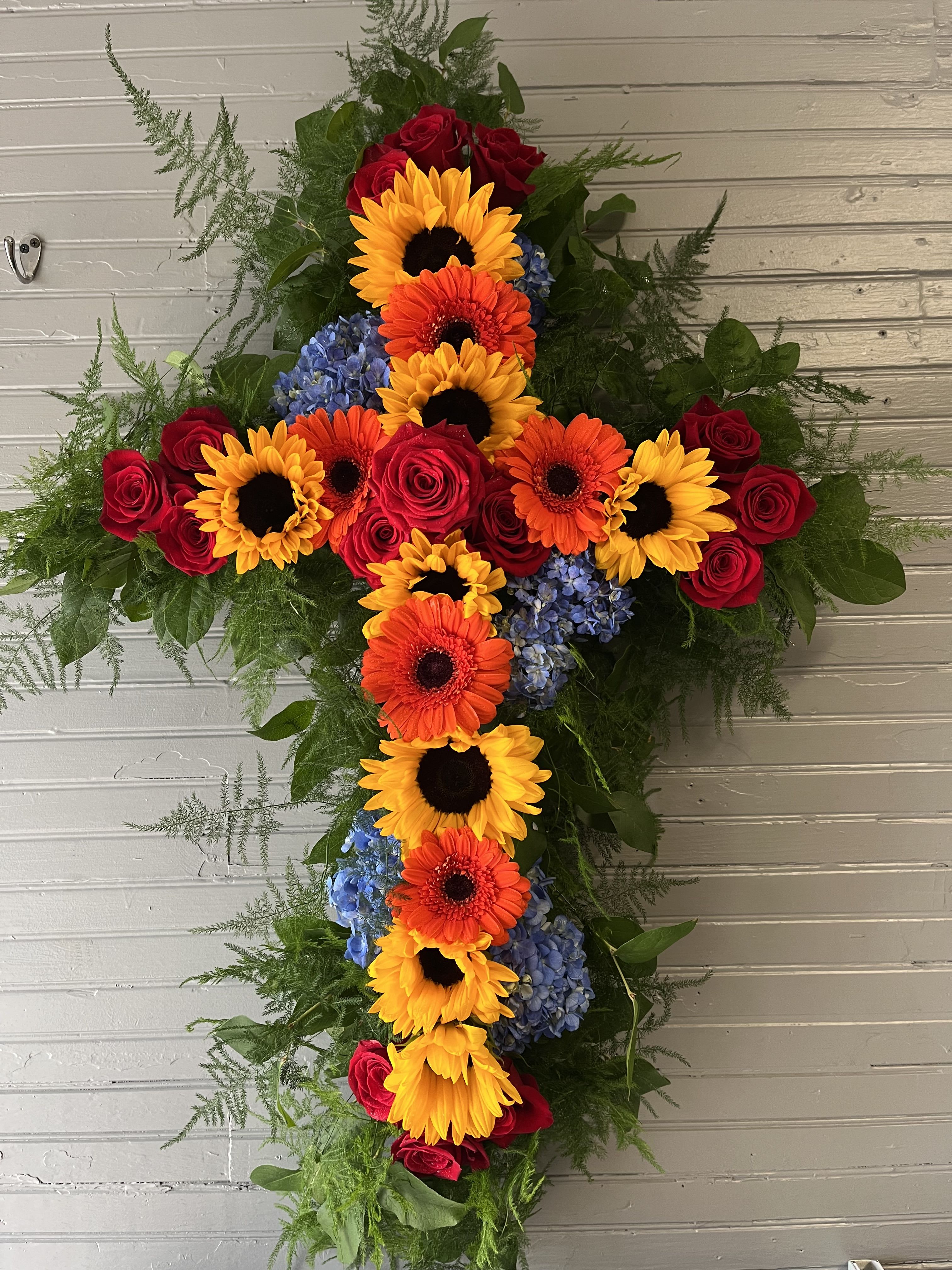 Cross-shaped floral arrangement with sunflowers, gerbera daisies, roses, and blue hydrangeas