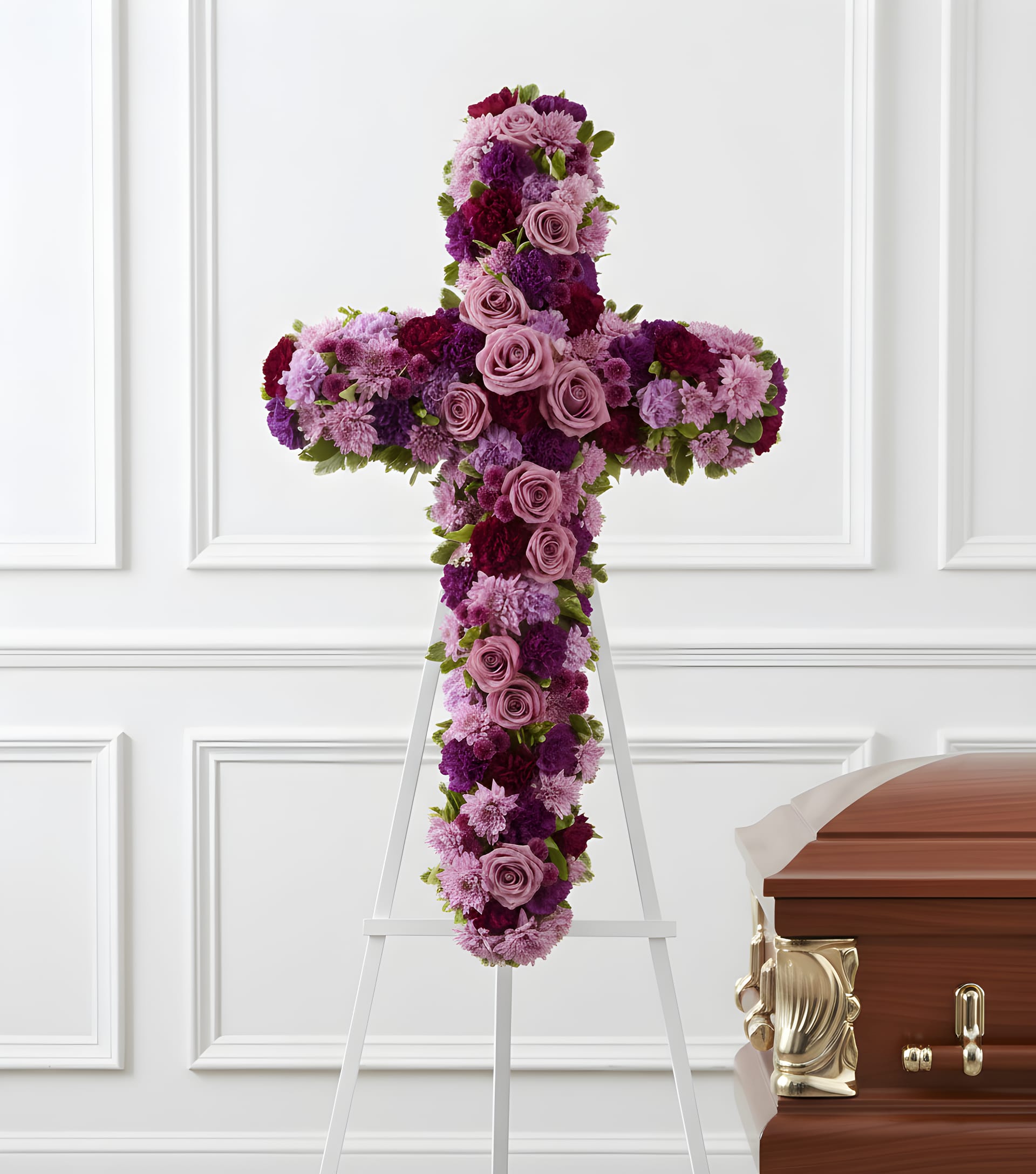 Purple and pink floral cross on a white easel beside a casket