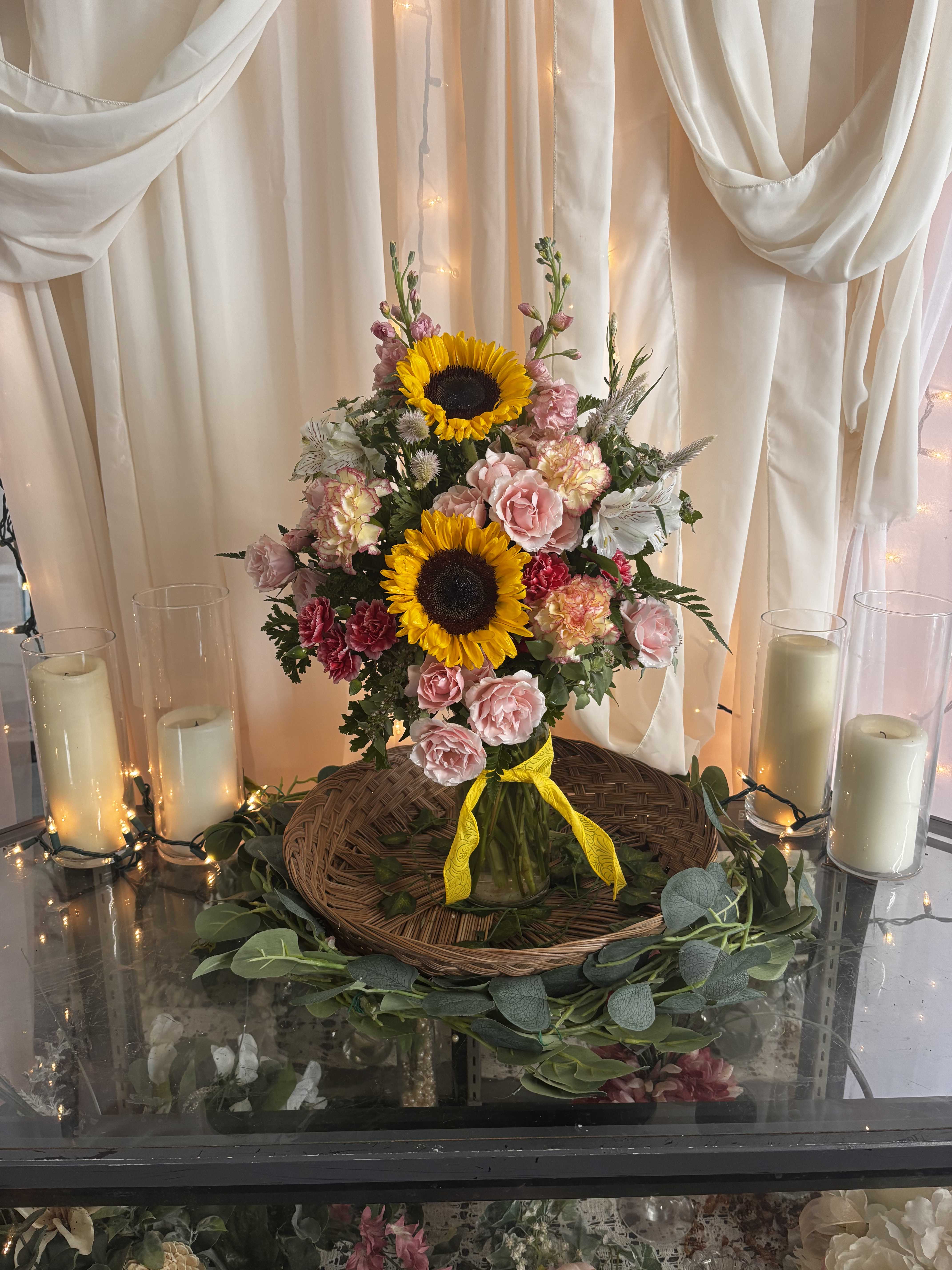 Sunflowers and pink roses arranged in a glass vase with candles nearby
