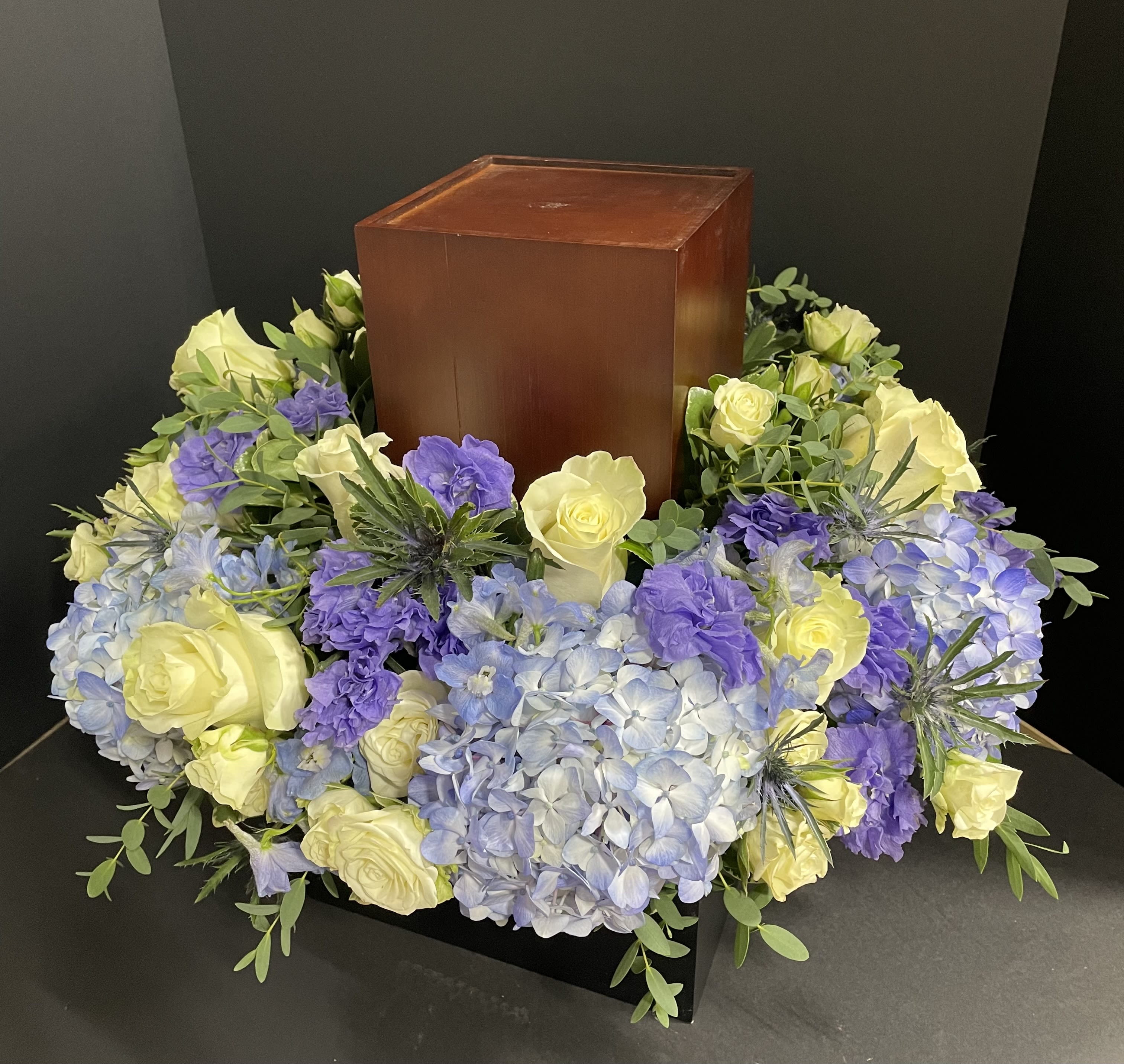 Floral wreath around a wooden urn with pale yellow roses and blue-purple hydrangeas