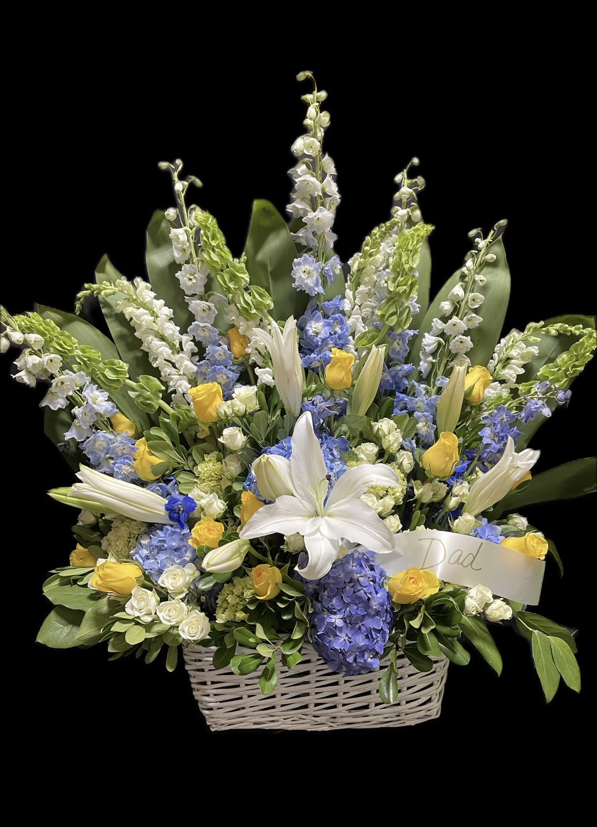 Basket arrangement of white lilies, yellow roses, and blue flowers with a Dad ribbon