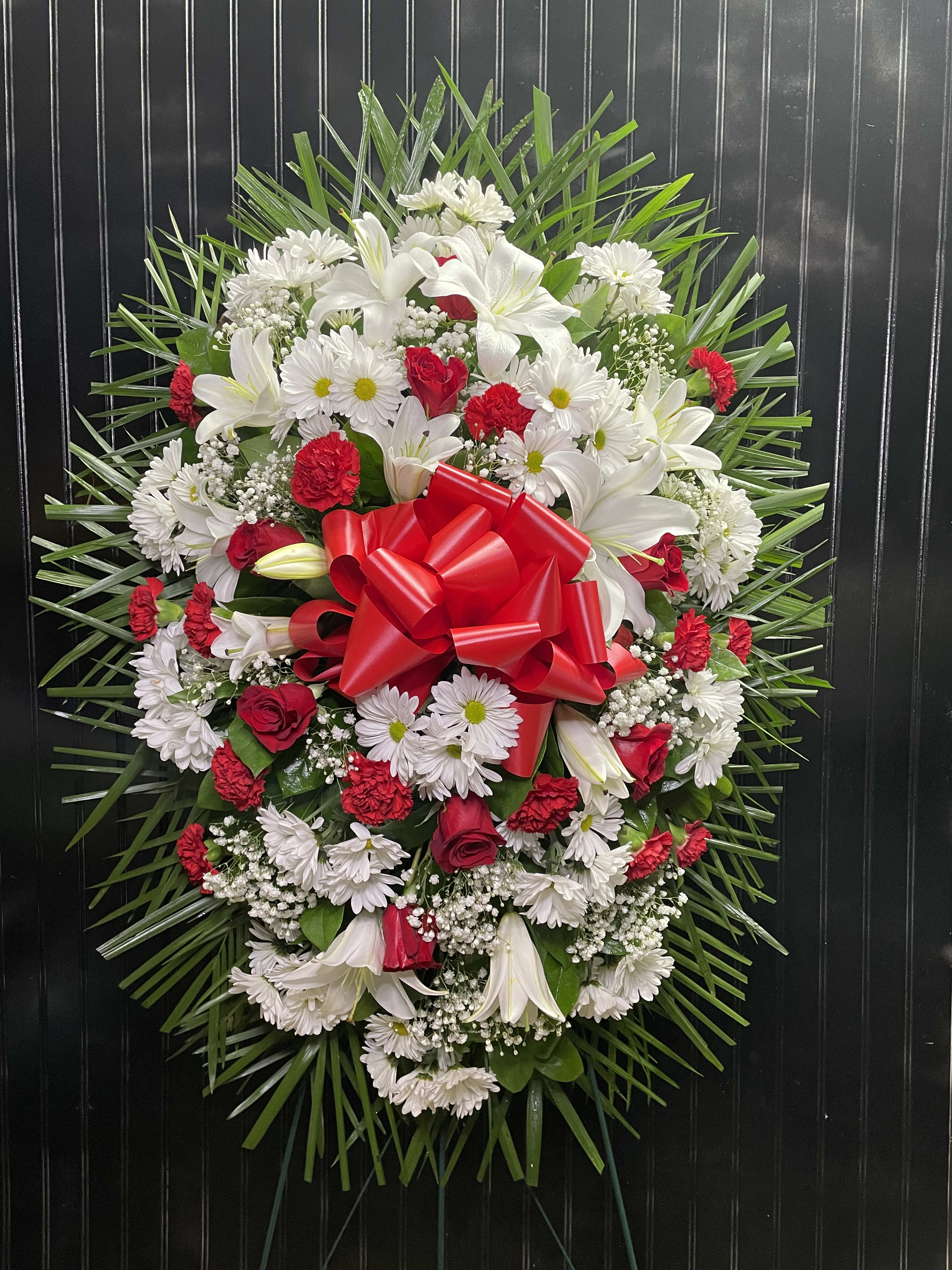 Oval floral wreath with white lilies, daisies, and red roses, finished with a red bow