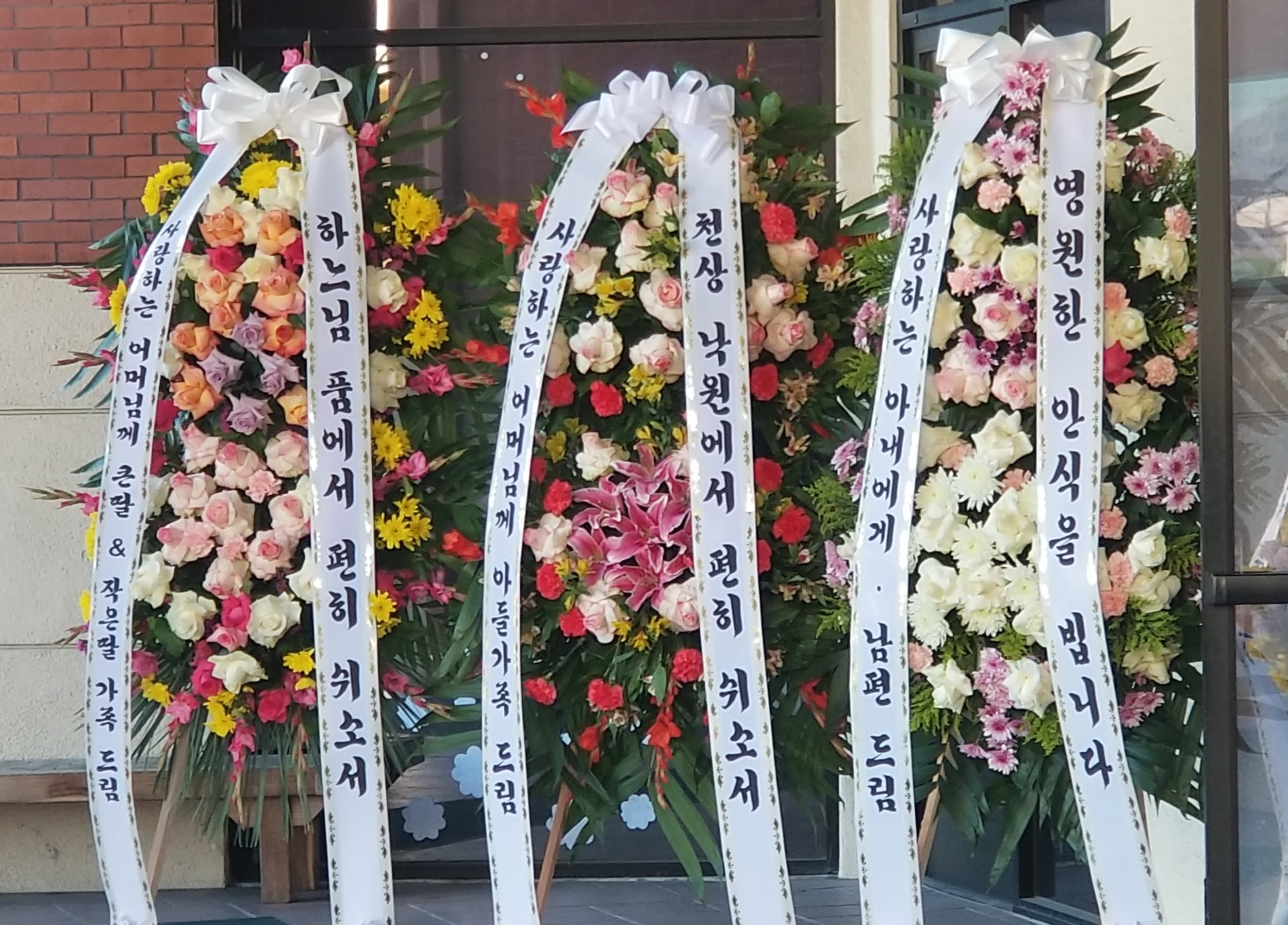 Three tall floral standing wreaths with white ribbon banners and mixed blooms