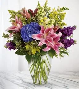 Bouquet of pink lilies, blue hydrangea, and purple flowers in a clear vase