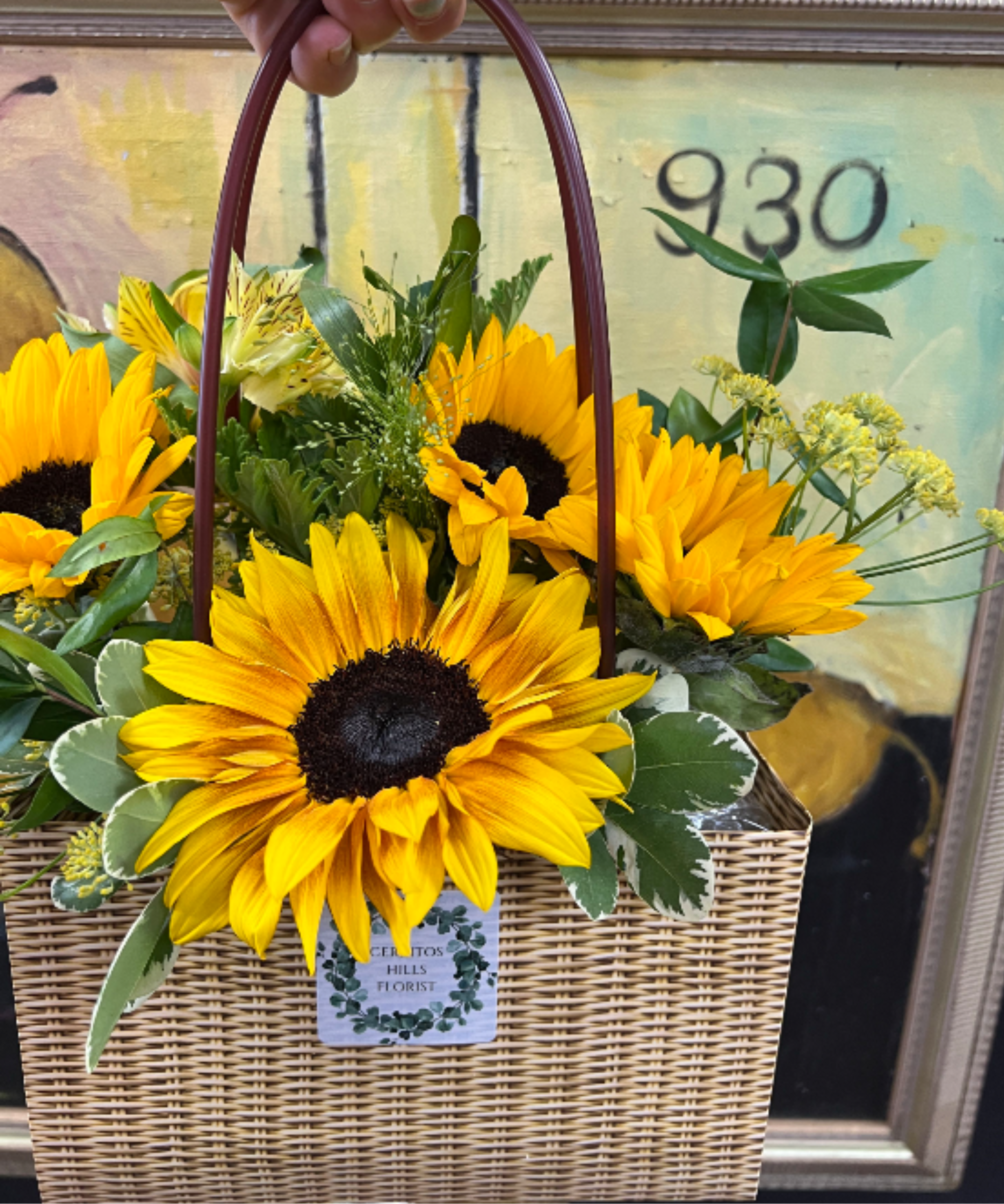 Father's Day flowers - Bright, hearty sunflowers take center stage in this Father's Day flower arrangement, a casual gift-basket bouquet that reads sunny, upbeat, and genuinely welcoming. Perfect as a Father's Day surprise, a birthday pick-me-up for dad, or a thank-you that says "you brighten my day," the yellow-and-green palette and rustic woven basket look presentation make the recipient feel noticed and appreciated.  Several large yellow sunflowers are clustered in a rectangular woven wicker basket with a burgundy arched handle, surrounded by mixed greenery and small pale filler blooms and pale yellow alstroemeria for texture and a layered, garden-like look. The arrangement sits low and open in the basket for an easy, homey display; a florist label and floral tag finish the presentation, making it ready to deliver straight to the person you're thinking of.  This arrangement is an everyday-style gift that brings cheer without fuss-ideal for dads who prefer casual, down-to-earth florals. No complicated care is needed: keep the basket in a cool spot away from direct sun and refresh water as needed to maintain the bright, cheerful blooms.