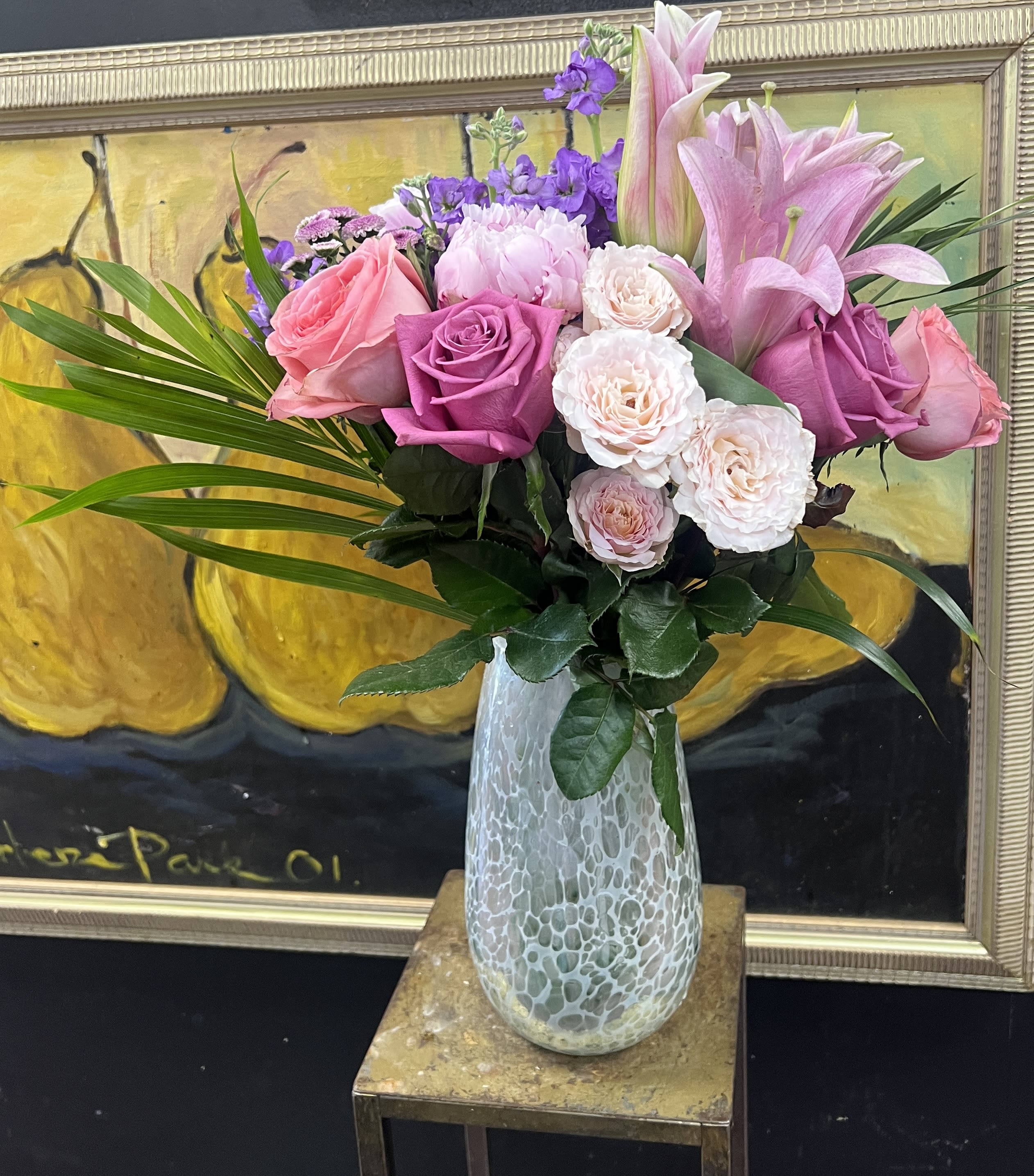 Happy Anniversary  - A lush cluster of pink, lavender and white roses crowned with tall pink lilies creates a romantic anniversary bouquet that also works beautifully for birthdays, thank-you gestures, or just-because surprises. The upright, abundant design reads celebratory and heartfelt-the person you're thinking of will feel genuinely seen when greeted with these soft, rosy tones and the whisper of purple accents.  This arrangement pairs full roses and showy lilies with small purple filler blooms and glossy rose foliage for texture and depth. Broad palm fronds fan the sides, adding dramatic width and green contrast, while a textured frosted glass vase gives the composition a polished, gift-ready finish.  Ideal for marking anniversaries, expressing love and romance, or saying thanks, this arrangement arrives ready to display; keep flowers in fresh water, trim stems every few days, and avoid direct sunlight to extend enjoyment. Select your size at checkout for the perfect presence-elegant enough for milestone moments, gentle enough for everyday celebration.