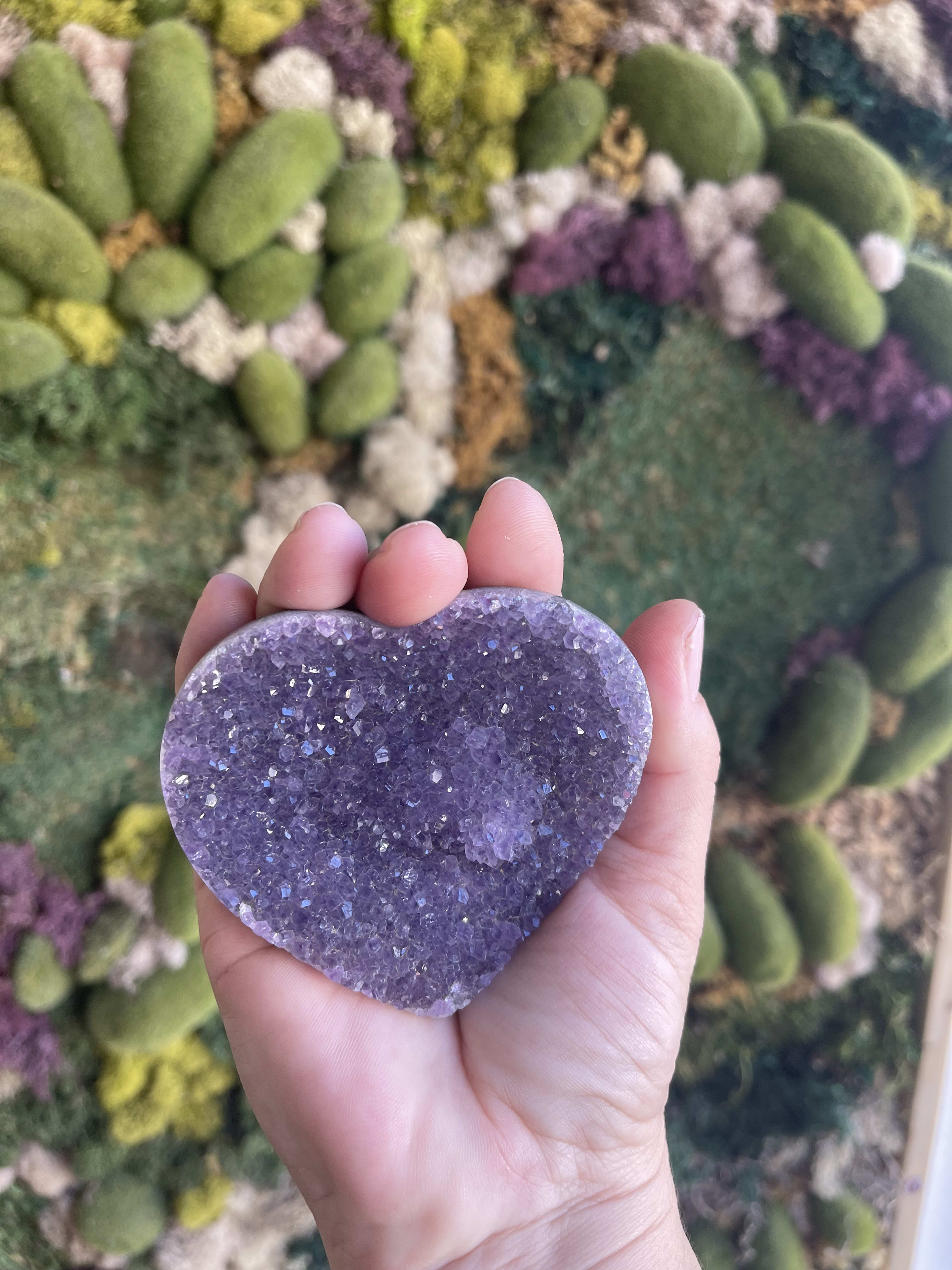 Hand holding a purple heart-shaped crystal against a floral wall
