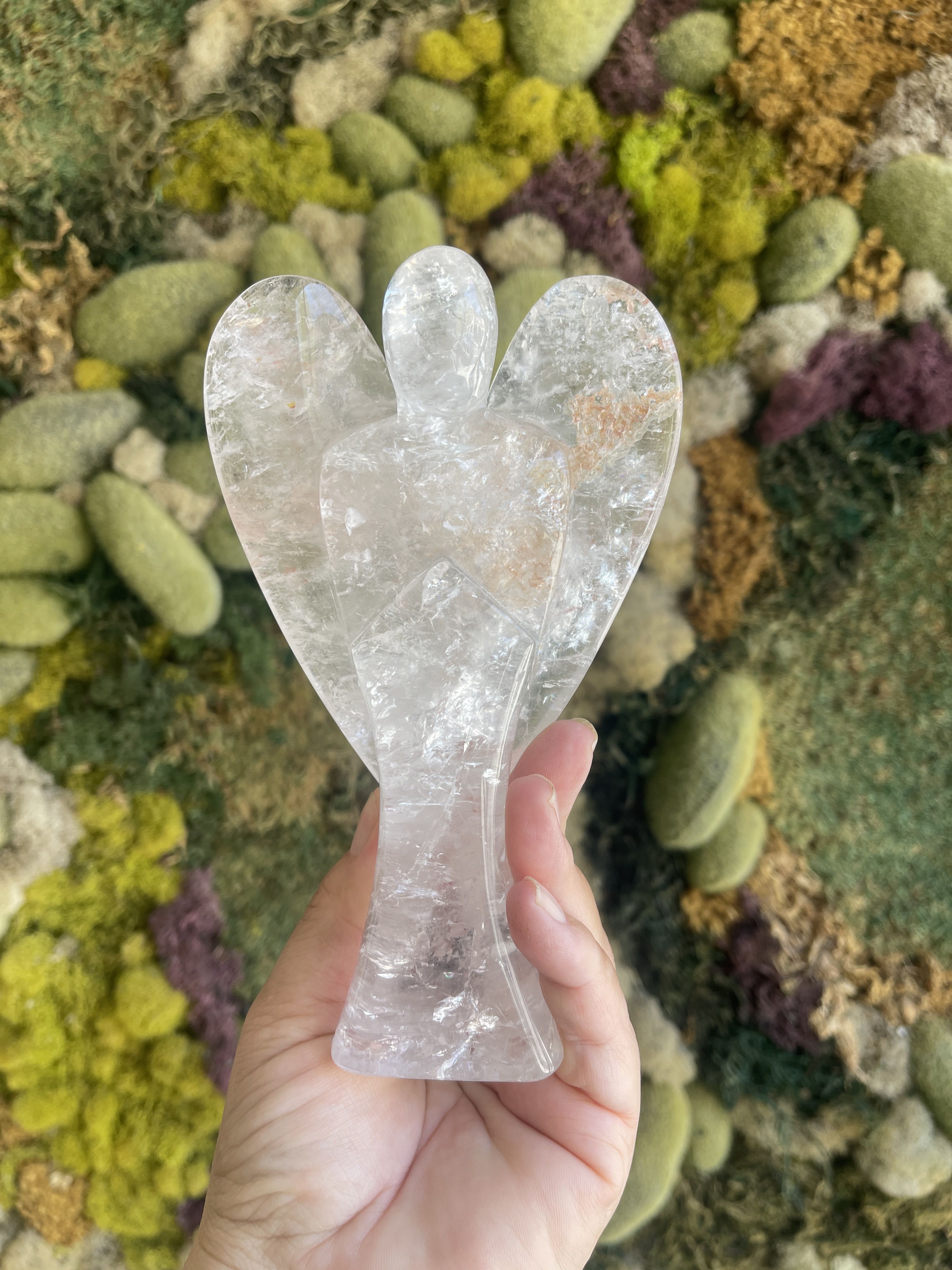 Hand holding a clear crystal angel figurine in front of a moss wall