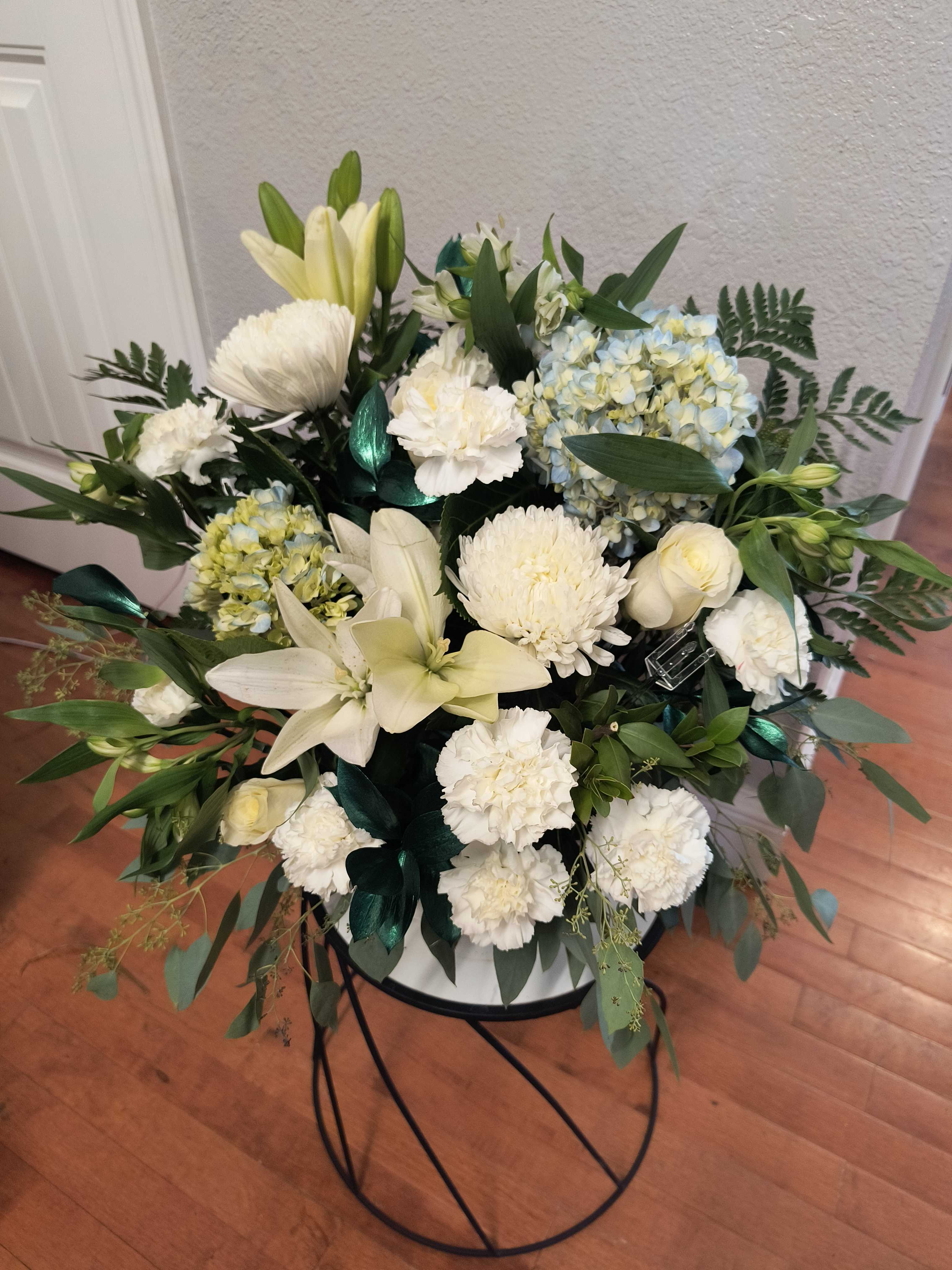 White floral arrangement with lilies, chrysanthemums, carnations, and blue hydrangeas in a stand