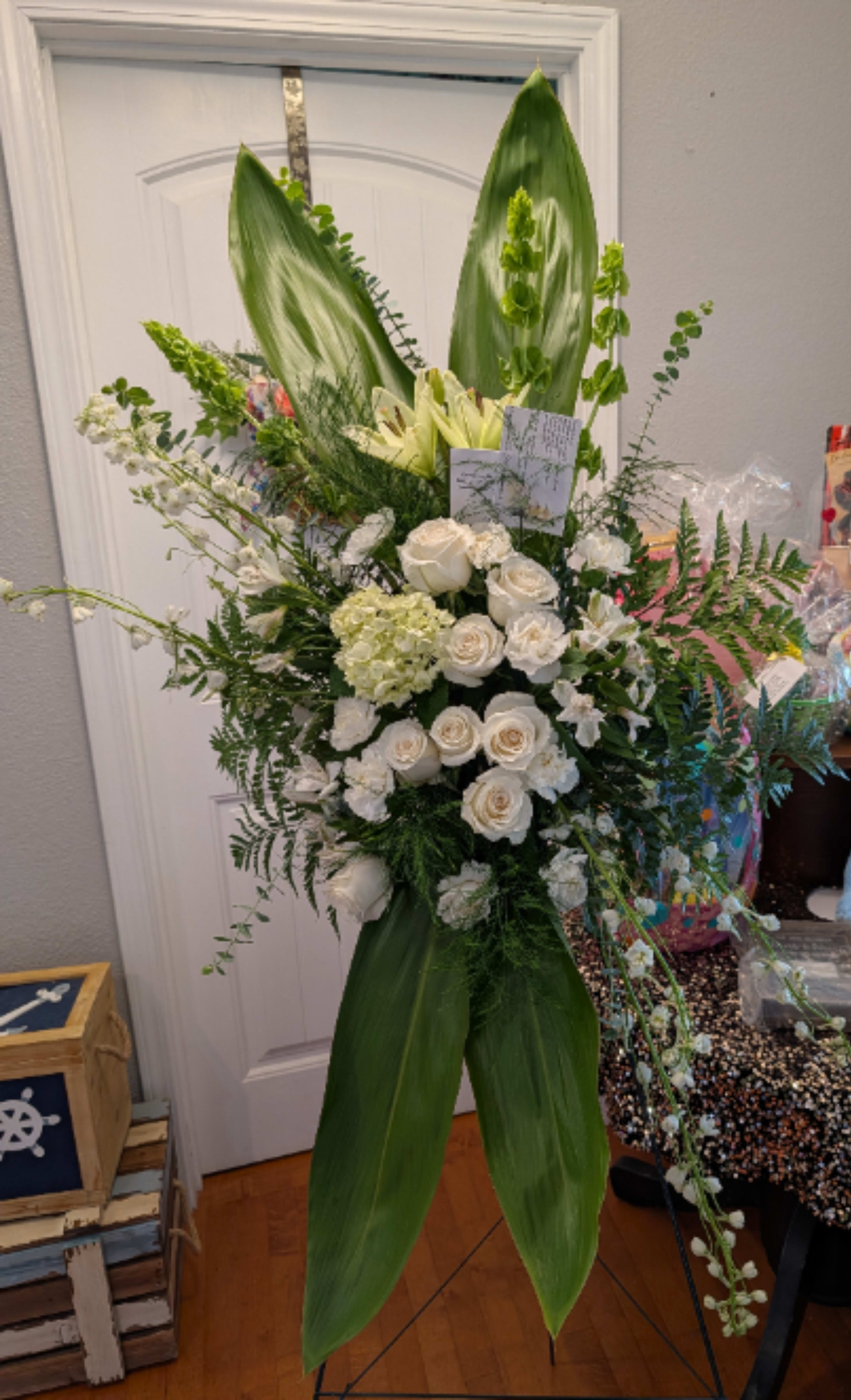 Heavenly Grace Cascade - This grand standing funeral spray features a highly formal, vertical architectural design that pairs crisp white garden blooms with dramatic, elongated tropical foliage. *Deluxe Size Shown*