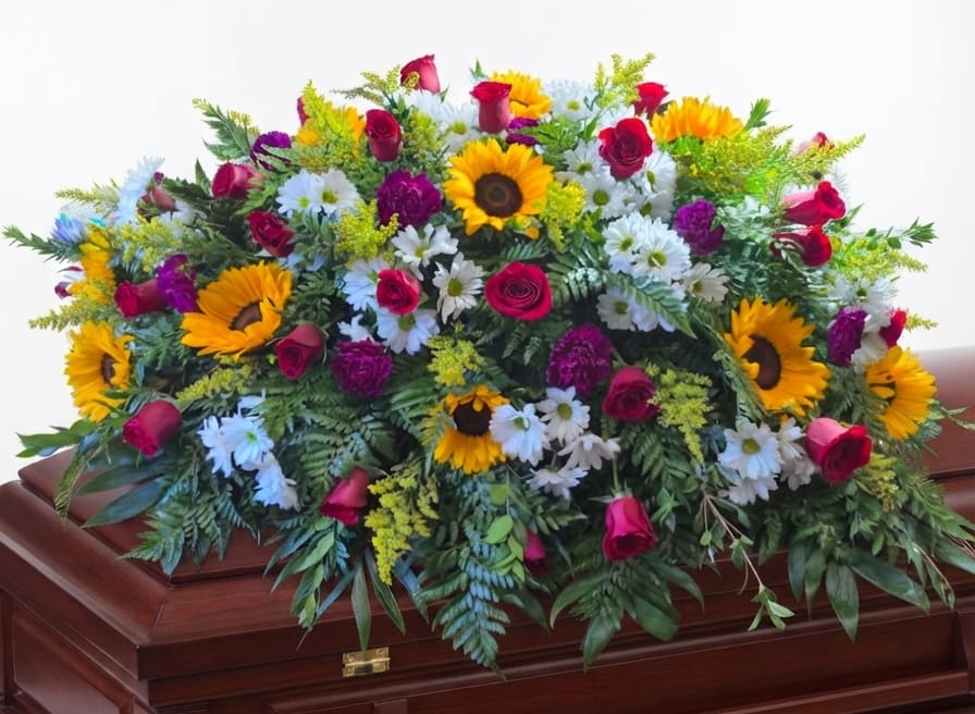 Large floral spray with sunflowers, roses, and white daisies on a wooden casket