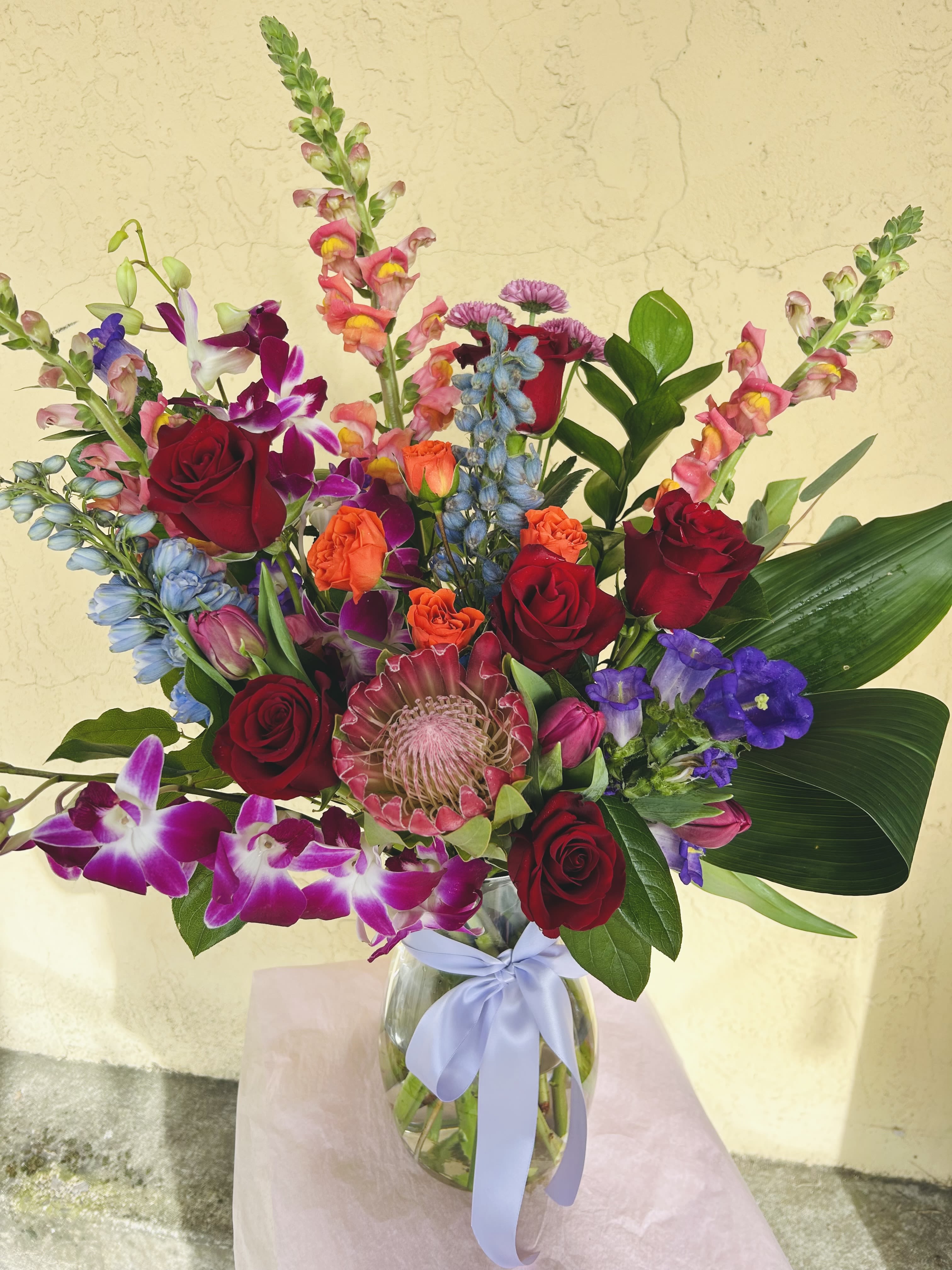 Mixed bouquet of red roses, orchids, and colorful blooms in a glass vase with ribbon