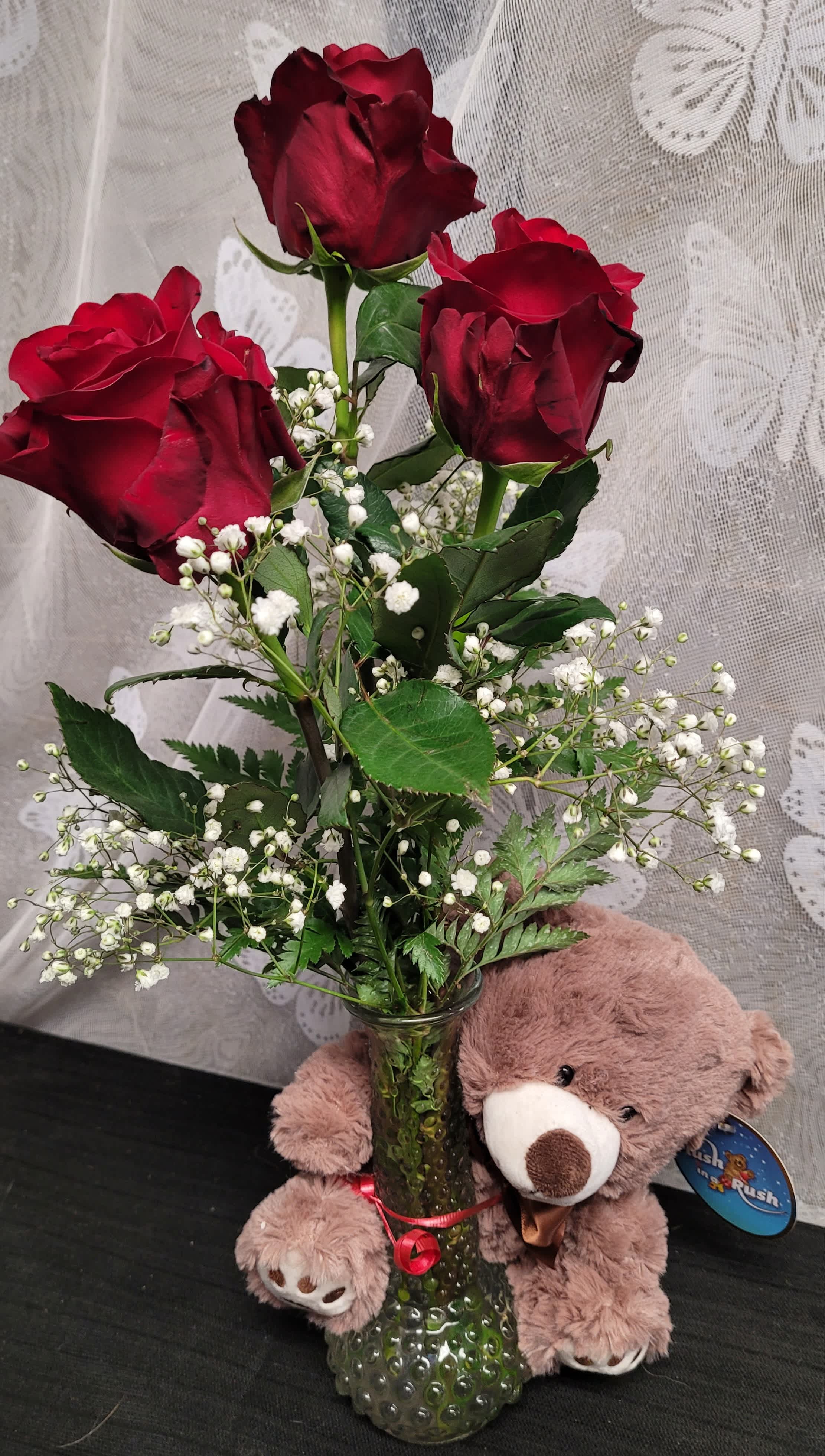 Three red roses in a glass vase with a teddy bear beside it