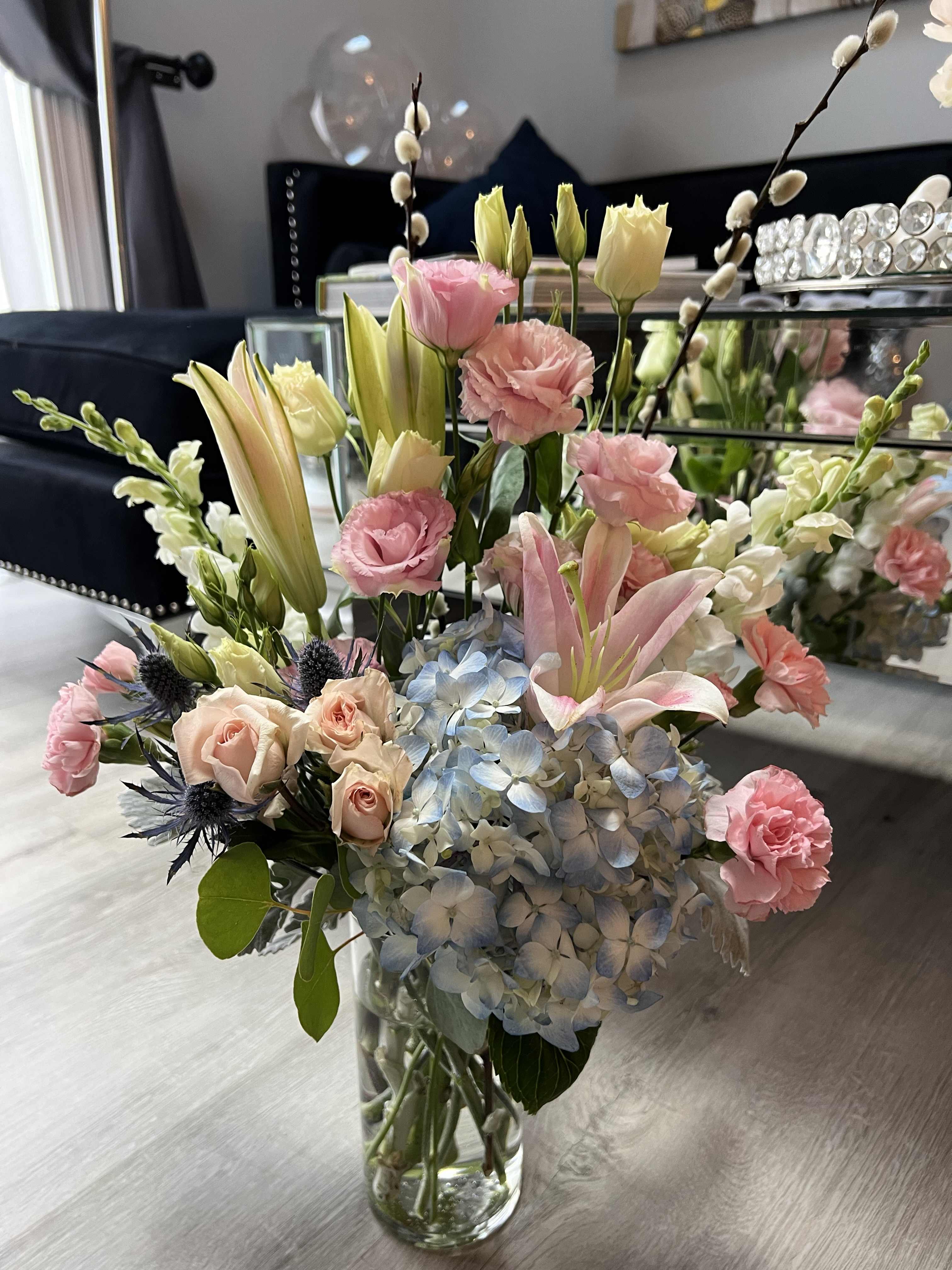 Mixed arrangement of pink lilies, roses, carnations, and blue hydrangea in a clear glass vase