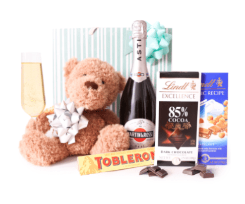 A TOAST - Soft tan plush and a sweet blush bow take center stage in this celebratory gift set designed for birthdays, congratulations, or a romantic toast. As a gift-friendly arrangement, A TOAST combines a cuddly teddy bear with a chilled bottle of sparkling wine and a single champagne flute to create an immediate mood of celebration-perfect for the person you want to make feel cherished and indulged.  Complementing the bear and bubbly are assorted chocolate bars, a candy box, and a coordinating gift bag, wrapped in a palette of pink, white, black, and bright blue that reads both playful and elegant. The pale striped backdrop and decorative bow give the whole composition a wrapped-gift presentation, making it ready to place on a table or bedside as a focal moment; textures range from the plush softness of the teddy to the glossy foil of the sweets for a tactile, indulgent experience.  This set works beautifully as a congratulations surprise, anniversary treat, or romantic pick-me-up-she'll feel celebrated, appreciated, and spoiled. Packaged for gifting, it arrives assembled and ready to present; no extra styling required.