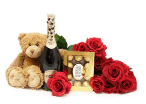 LOVE PACKAGE - A cluster of vivid red roses takes center stage in this romantic flower arrangement, paired with a plush teddy bear, a bottle of champagne, and a boxed assortment of chocolates-perfect for a Valentine's Day or anniversary gift. The bold red blooms create an immediate impression of passion and celebration, while soft tan and gold accents from the bear and packaging add warmth and luxury that will make the person you're thinking of feel truly cherished.  This compact gift set focuses on roses as the floral element, surrounded by indulgent complementary items rather than a full traditional bouquet. The arrangement reads as a mixed floral-and-gift package: bright red roses for romance, a cuddly teddy for comfort, premium chocolates for sweetness, and champagne to toast the moment. It's an easy, thoughtful way to mark anniversaries, romantic milestones, or any occasion when you want to say "I love you" with both flowers and a keepsake.