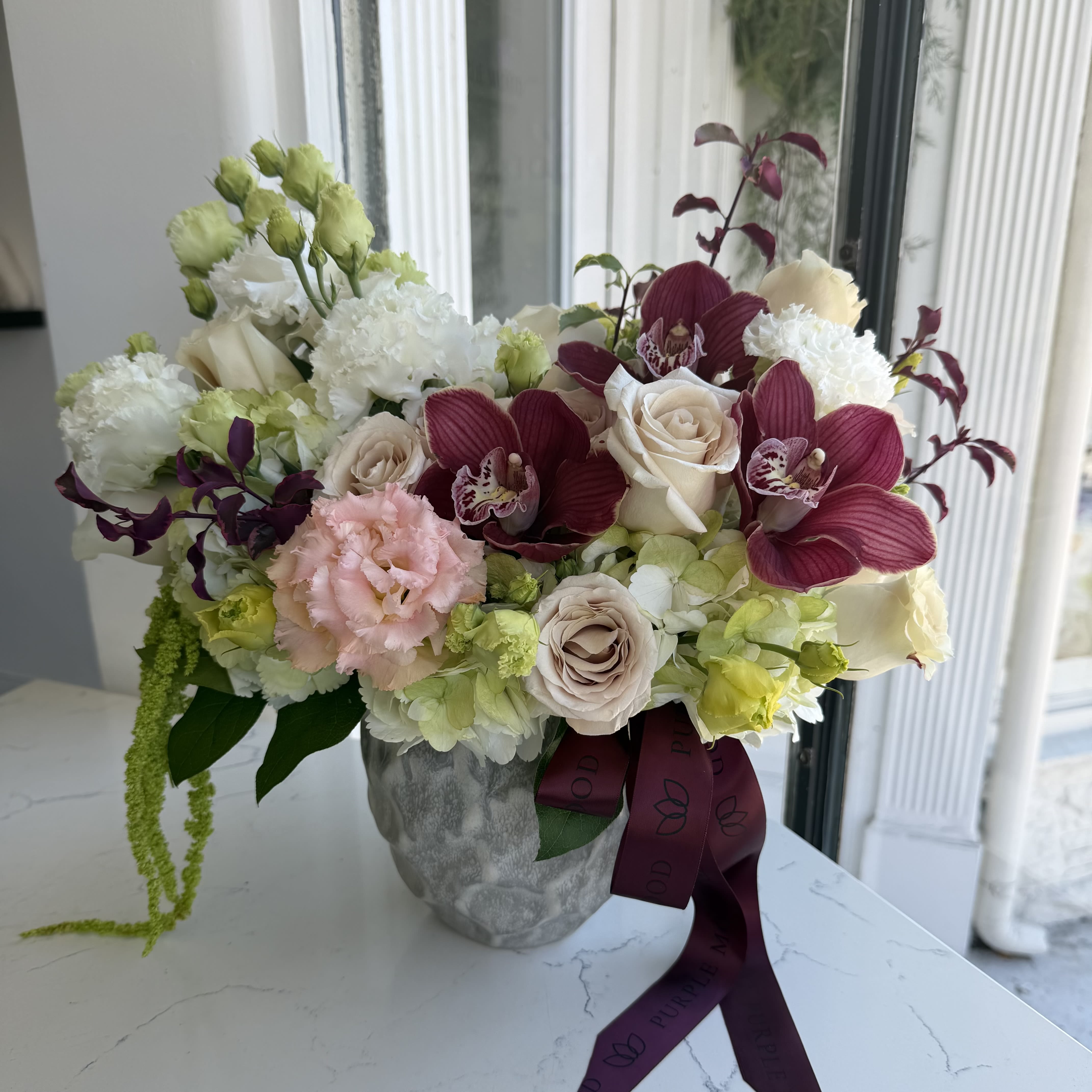 Orchid elegance Vase - A refined and elegant arrangement featuring premium orchids, blush roses, ranunculus, and seasonal blooms in soft ivory and burgundy tones. Designed in a modern stone vase, this piece brings a perfect balance of softness and richness, making it ideal for any occasion.  This arrangement is thoughtfully styled with a natural, airy feel while maintaining a full and luxurious look.  Please note: • Vase is included • Flowers may vary slightly based on availability while maintaining the overall color palette and style • Perfect for birthdays, celebrations, or as a sophisticated everyday gift
