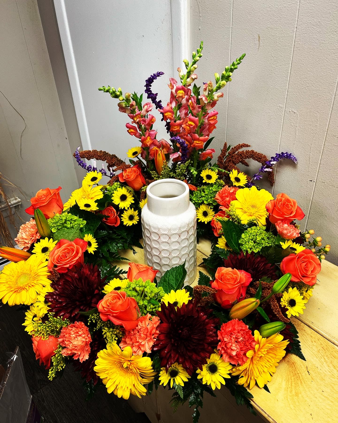Circular floral arrangement with orange roses, yellow daisies, and a white vase in the center
