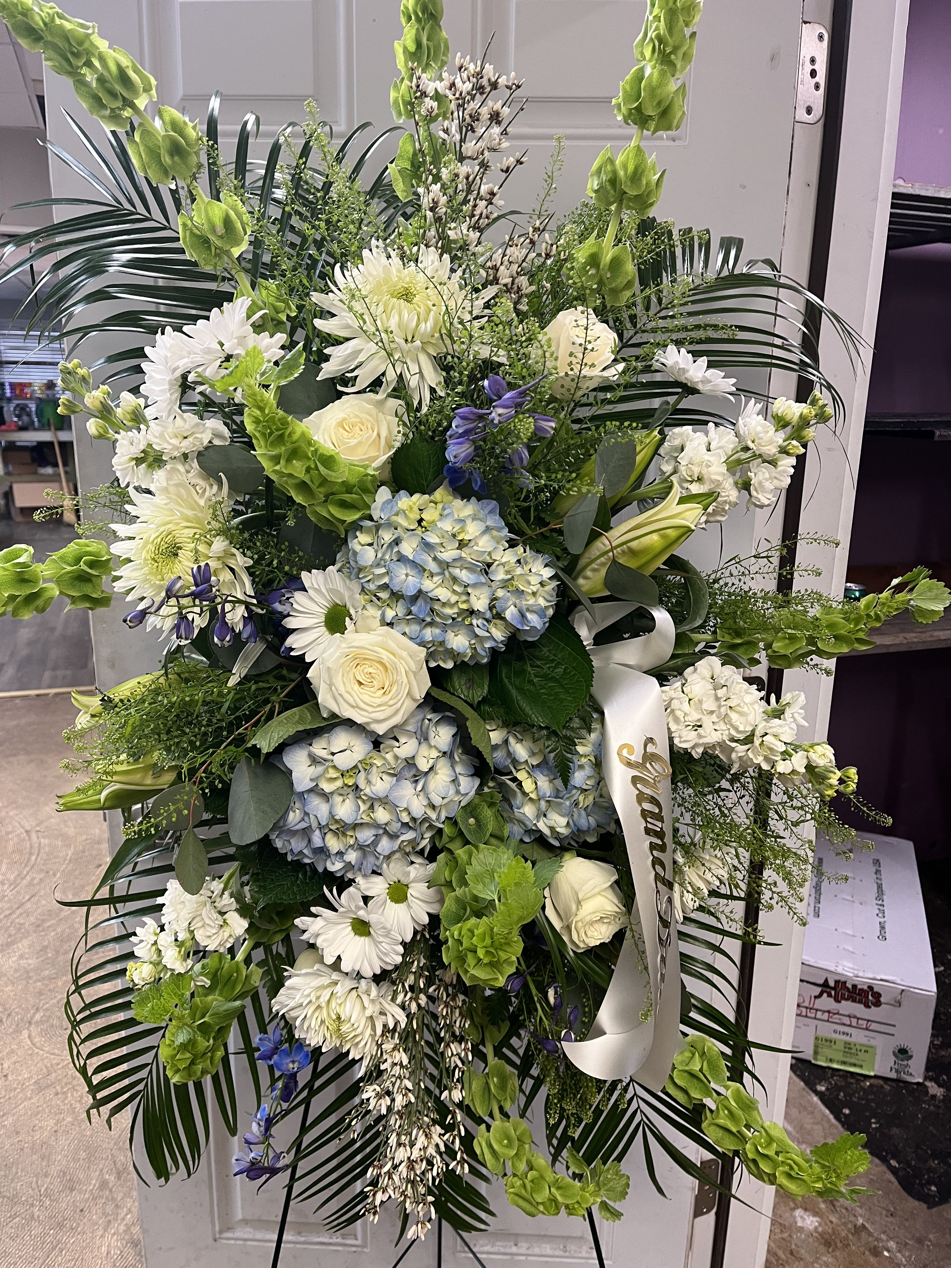 Blue, white and green standing spray - Designers choice of blue, white and green flowers arranged in a standing spray. Perfect statement piece for a sympathy arrangement. Script on banner can be customized.