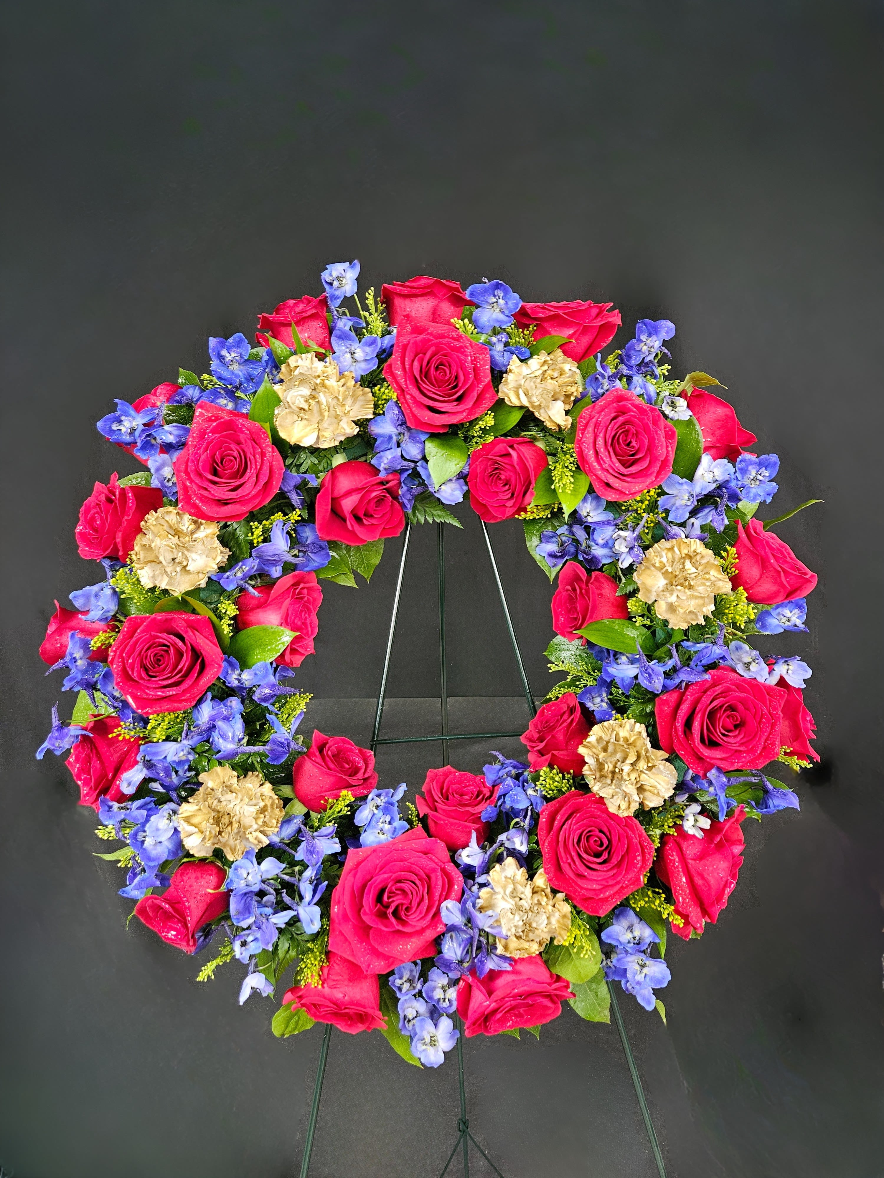 Heart-shaped wreath of pink roses, blue flowers, and gold carnations on a stand