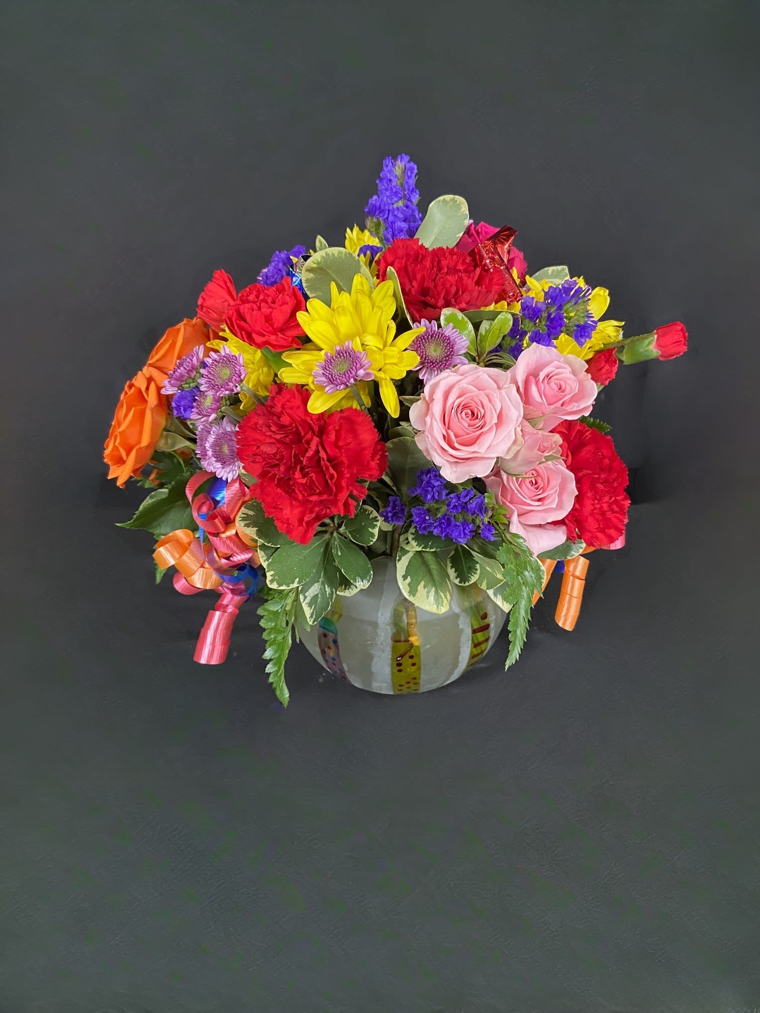 Brilliant Birthday Blooms - A frosted globe is decorated with birthday candles and filled with seasonal flowers such as yellow daisies, orange roses, red carnations, and multi color alstro. Celebratory streamers trail out of the sides with party favors peaking out the top.