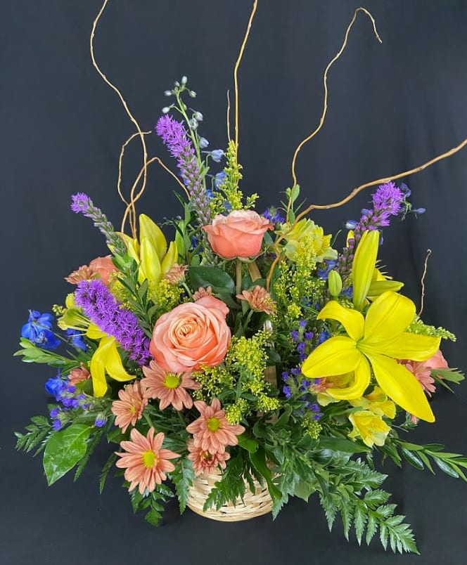 Basket arrangement with peach roses, yellow lilies, purple spikes, and blue flowers.