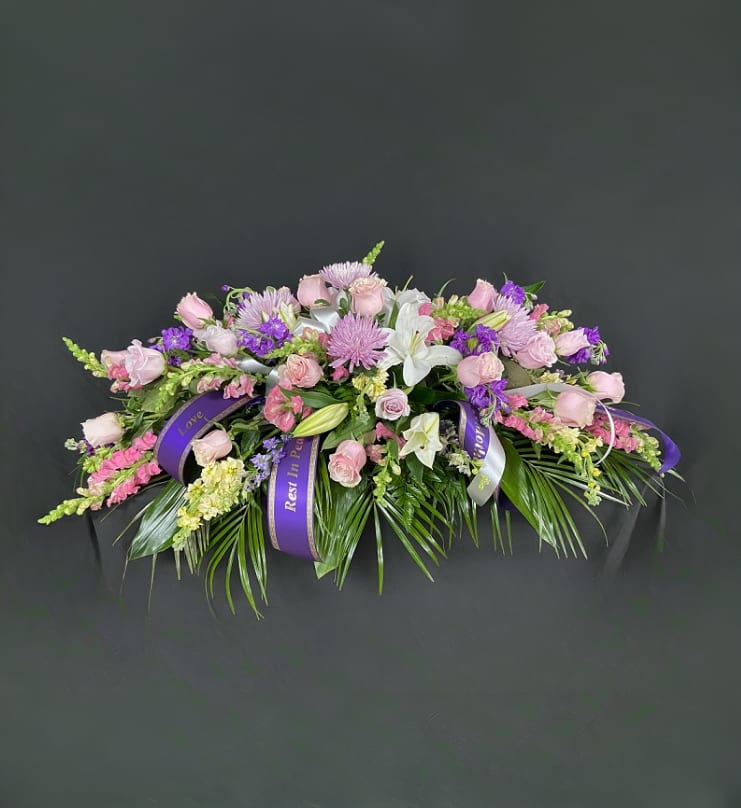 Pink and purple floral casket spray with white lilies and ribbon