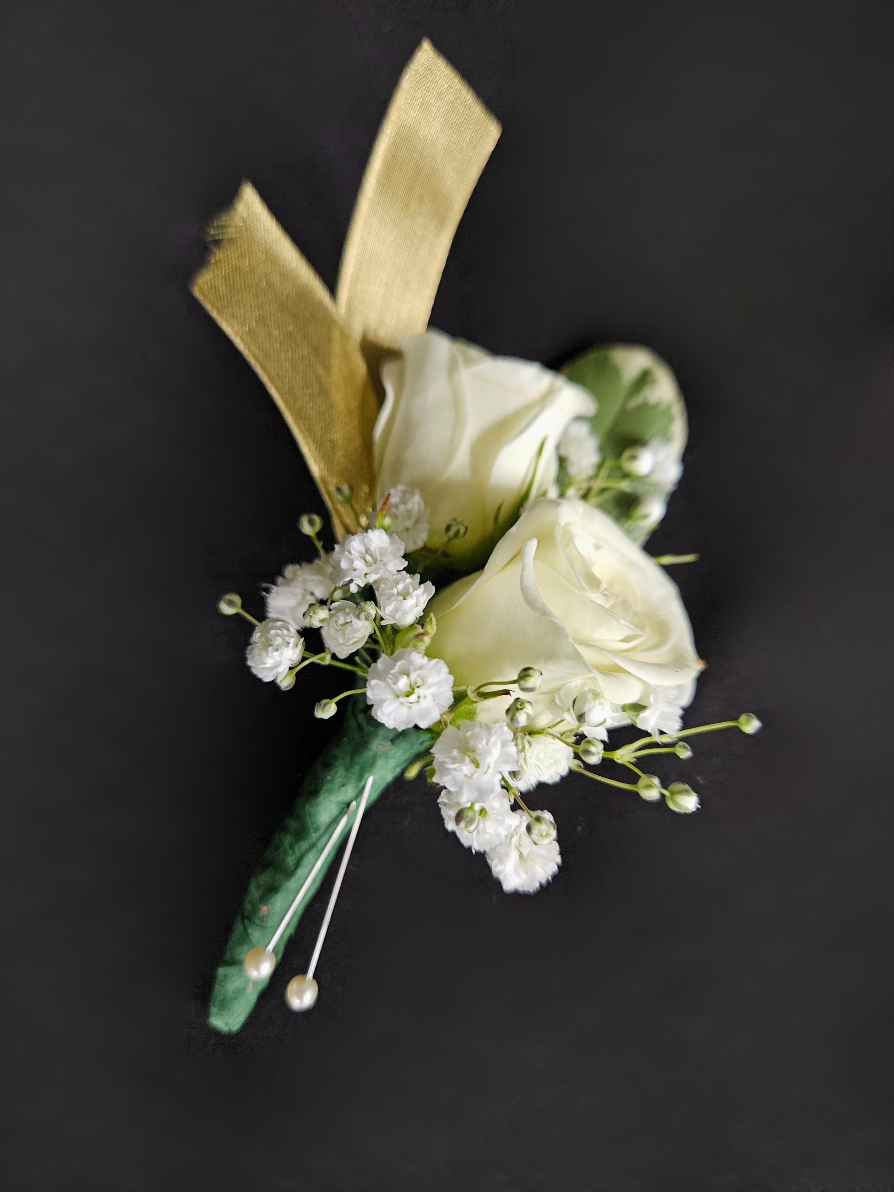 White rose boutonniere with tiny white filler flowers, gold ribbon, and pearl pins on a dark background.