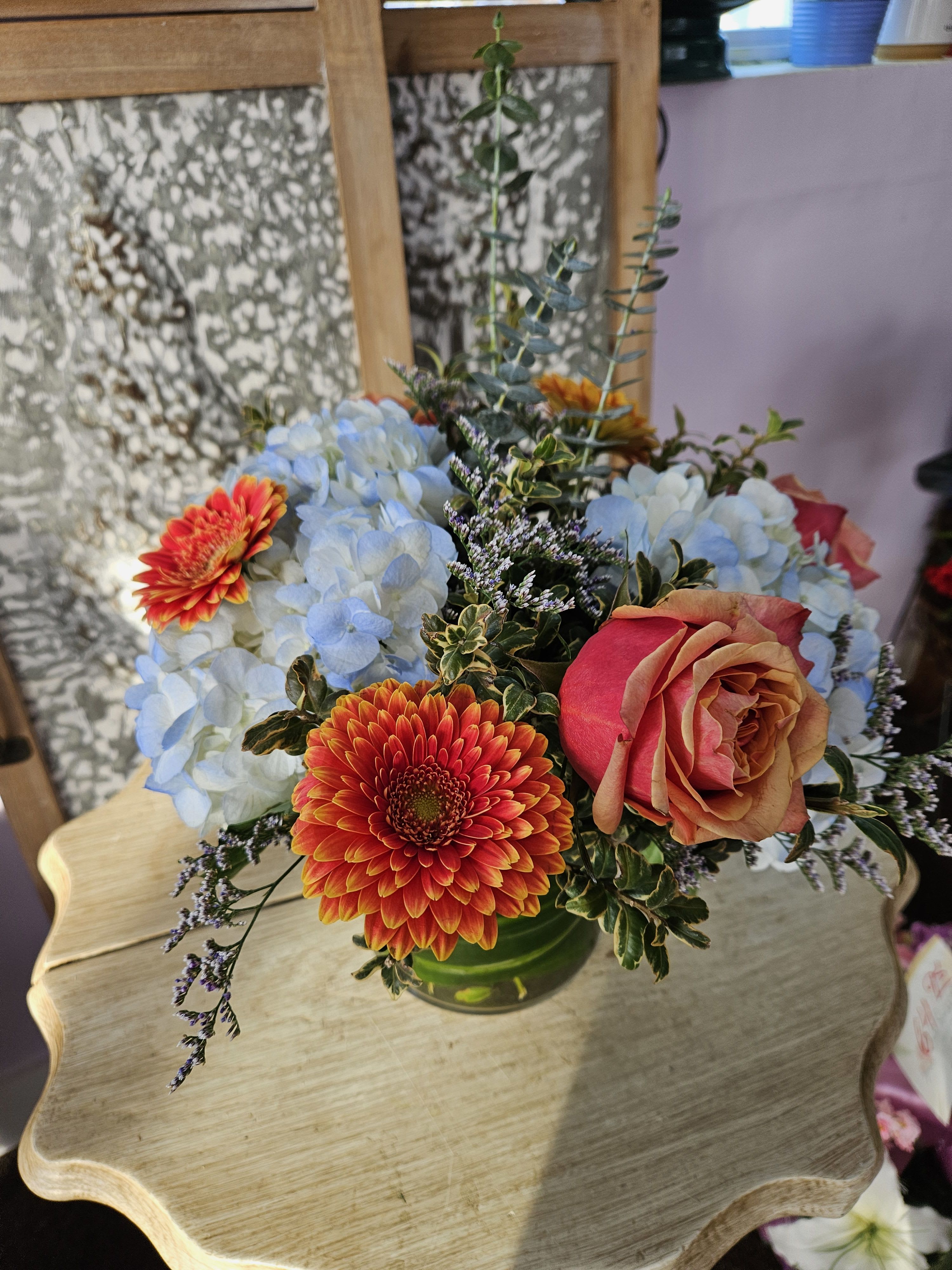 Low arrangement of orange gerbera, peach rose, and blue hydrangeas in a clear glass vase.