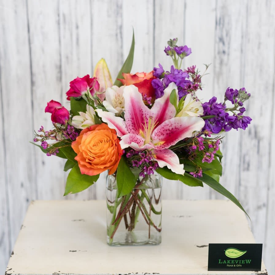 Popsicle - A vibrant cluster of color and texture, Popsicle is a cheerful mixed flower arrangement perfect as a birthday surprise, or a just-because pick-me-up. The compact floral design centers on a large, locally grown, stargazer lily surrounded by sunset roses, magenta spray roses, and purple filler blooms, creating an energetic, summery palette that reads as both playful and sophisticated. This is the kind of arrangement that makes the person you're thinking of feel seen and celebrated-whether you're saying "thank you," "get well soon," or "I'm sorry."  Arranged in a clear square glass vase, the composition is full and layered with blooms spreading outward in a lively, slightly asymmetrical shape that suggests a fresh garden bouquet. Stock and other textured fillers add scent and visual depth while the combination of pink, purple, white, and orange keeps the look bright and approachable.   Approximate dimensions: 12" H x 12" W