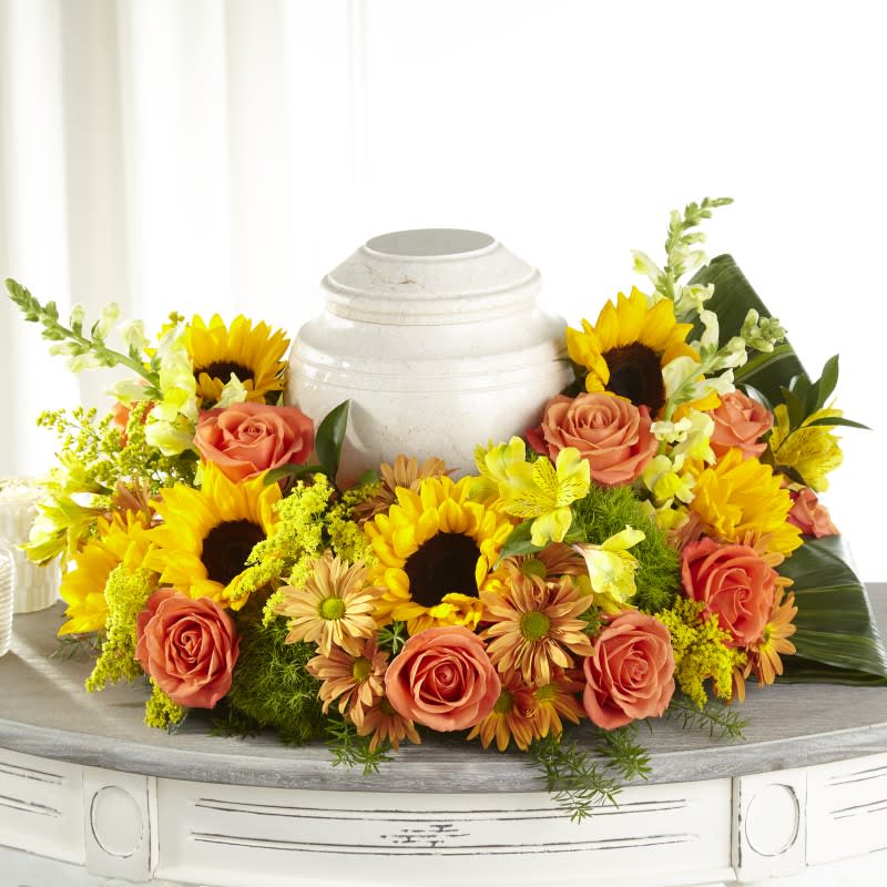 Loving Embrace Cremation Adornment  - Loving Embrace Cremation Adornment offers a comforting display to accent an urn or box, framed in the center of a ring of beautiful blooms. This beautiful ring of vibrant yellow and orange flowers is handcrafted with sunflowers, roses and pompons to celebrate their beautiful life.  