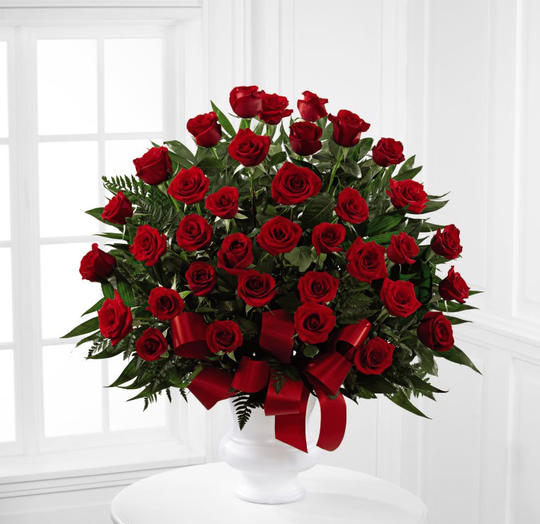 Blessed With Love - A rich display of the love shared throughout the life of the deceased. Three dozen brilliant red roses are elegantly displayed in a white urn and accented with lush greens and red satin ribbon to create a beautiful tribute to honor your special relationship. (Other color roses available upon request.)  S15-4470S