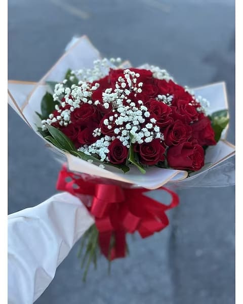 Handheld bouquet of red roses with white filler flowers wrapped in paper and tied with a large red bow.