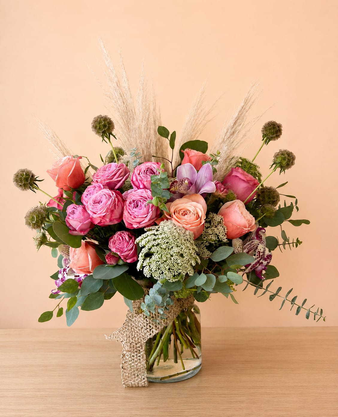 Beach Vibe - Bring the serene elegance of the coast indoors with this unique beach-inspired floral creation.   Artfully arranged in a clear glass cylinder wrapped with a natural tan burlap ribbon, this design beautifully balances rustic texture with refined blooms.  Displaying lush garden roses, exotic cymbidium &amp; phalaenopsis orchids, accented with airy green mist and feathery pampas grass. While scabiosa pods and lush eucalyptus add organic depth &amp; movement.  Perfect for coastal weddings, summer celebrations, or anyone dreaming of salty air and endless horizons, this arrangement captures the effortless beauty of the beach with a sophisticated twist.