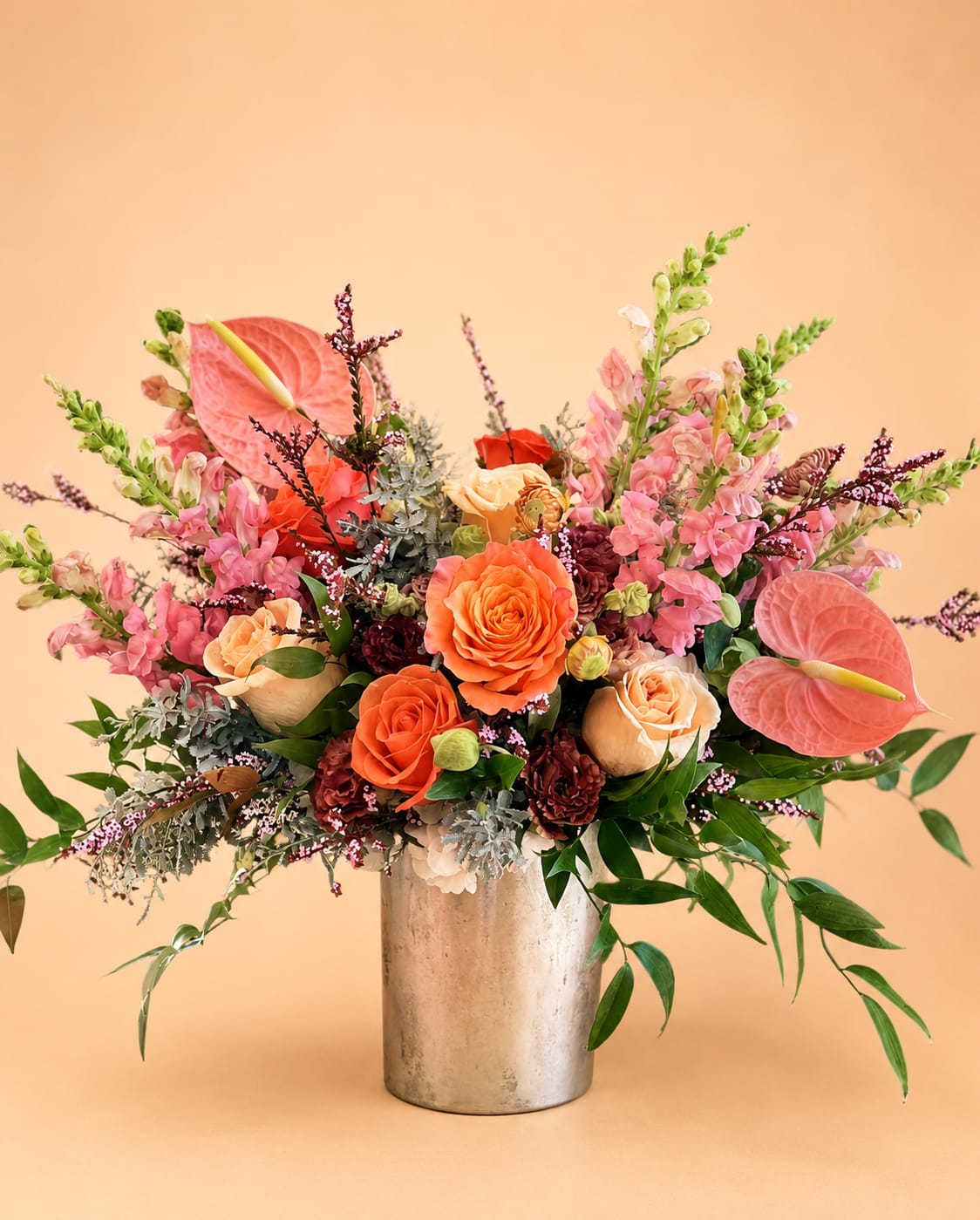 Cleo Sol  - Our "Cleo Sol" arrangement is a refreshing way to celebrate bright sunny days. This arrangement captures the essence of the sun in an energetic and zesty way.   Florals included: Free Spirit roses, pink anthuriums, pink snapdragons, orange crush roses, mauve lisianthus, pink scottish heathers, italian ruscus, and acacia.  Designed in a silver 5x5 cylinder vase. This arrangement stands approximately 15" inches tall and 22" inches wide. 