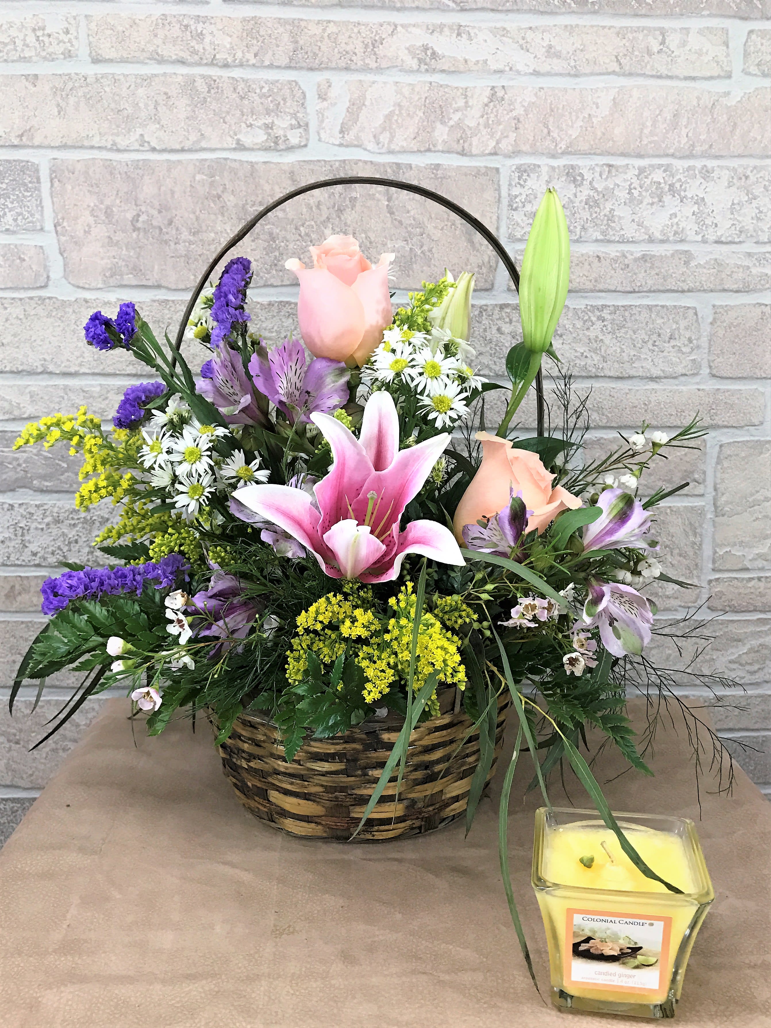 Basket arrangement with pink lilies, roses, and purple flowers beside a yellow candle