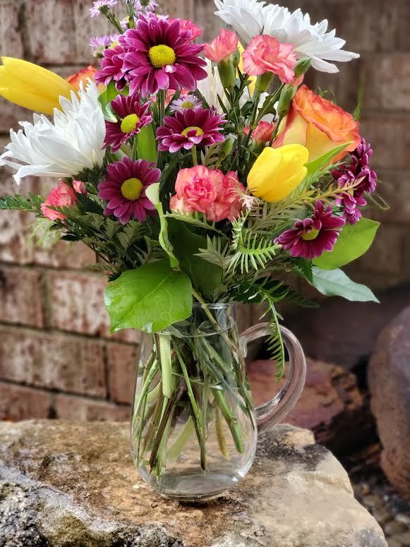 Mixed bouquet of daisies, tulips, and carnations in a clear glass pitcher
