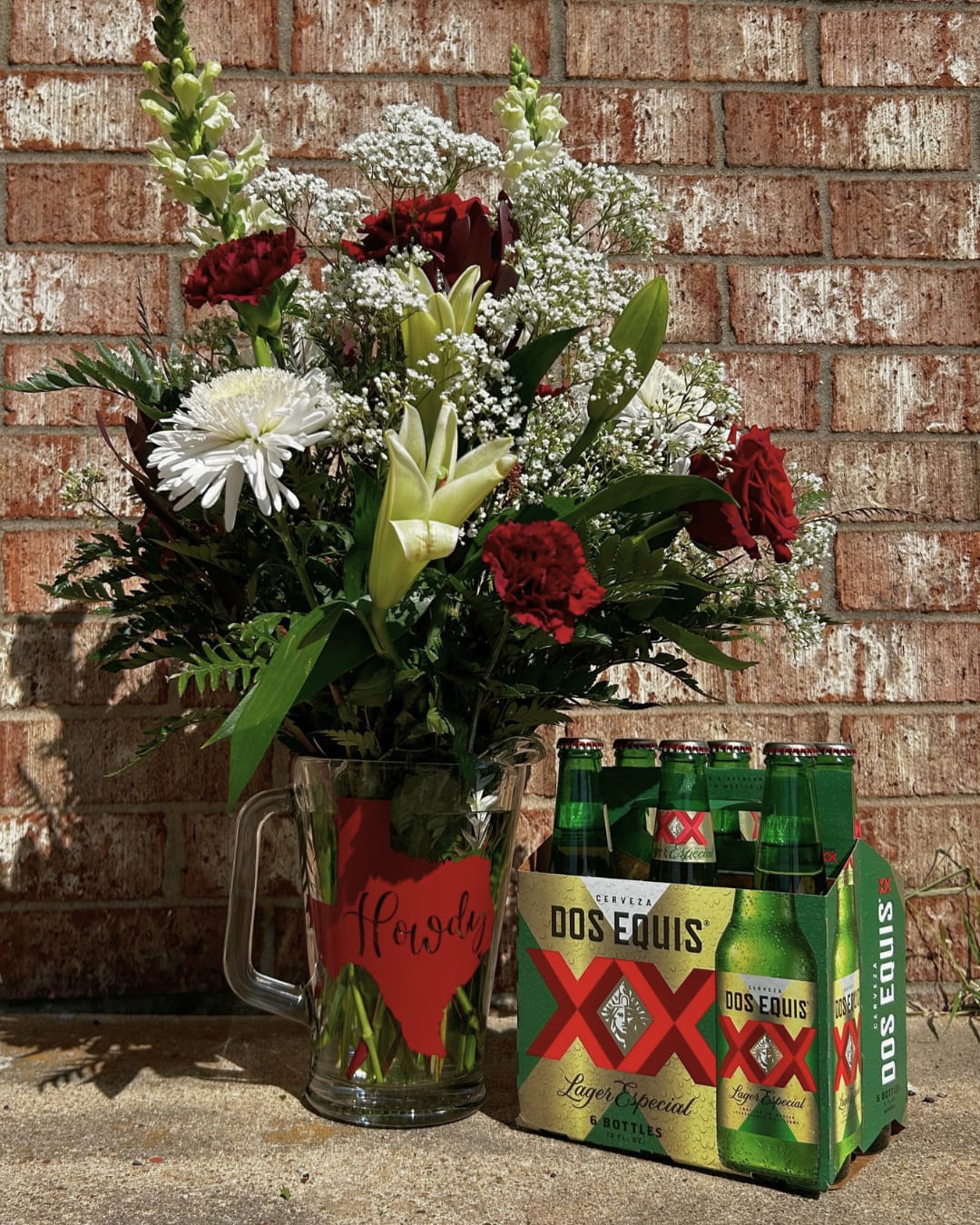Mixed flower arrangement in a glass mug beside a six-pack of Dos Equis beer