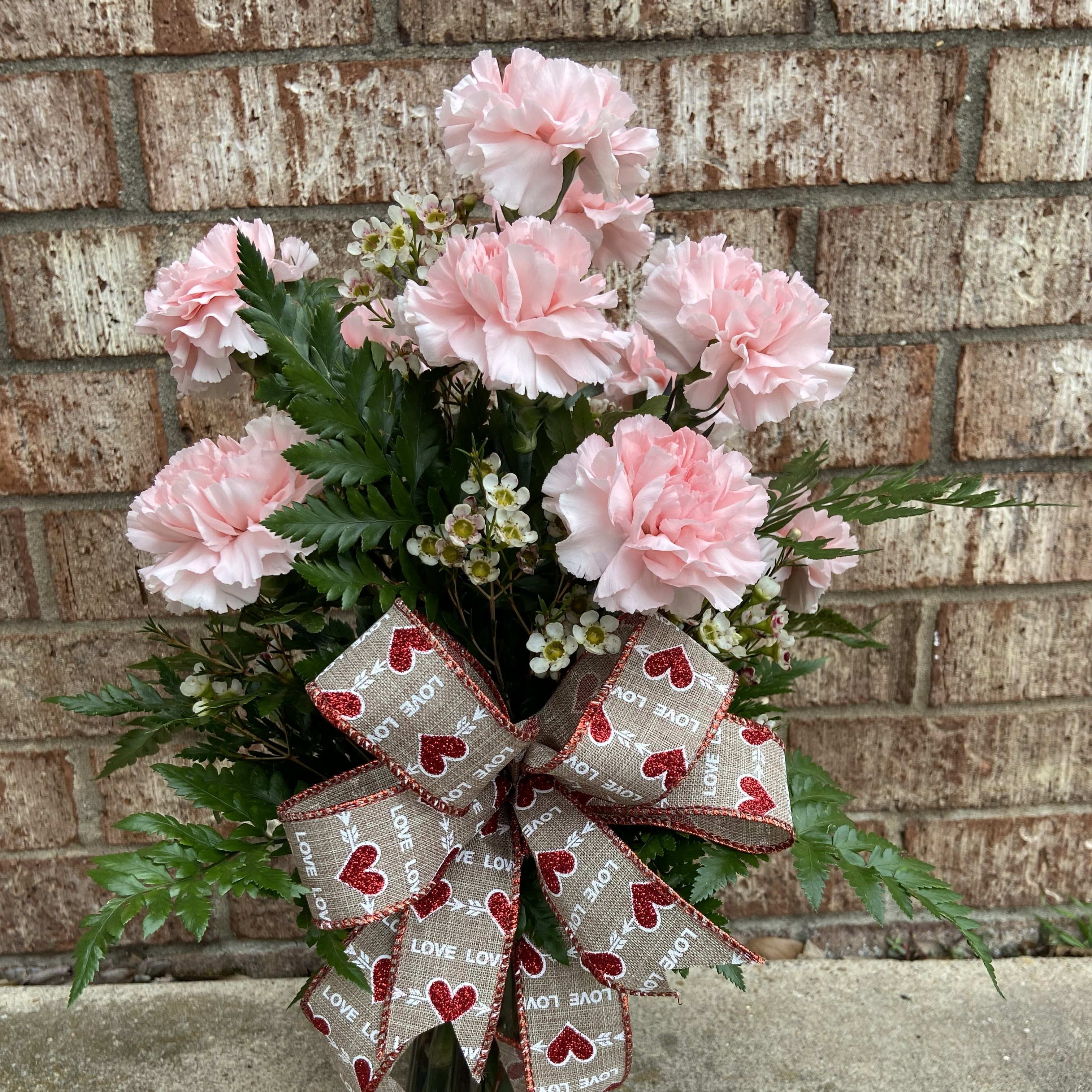 Pink carnations arranged with greenery and a heart-patterned ribbon bow.