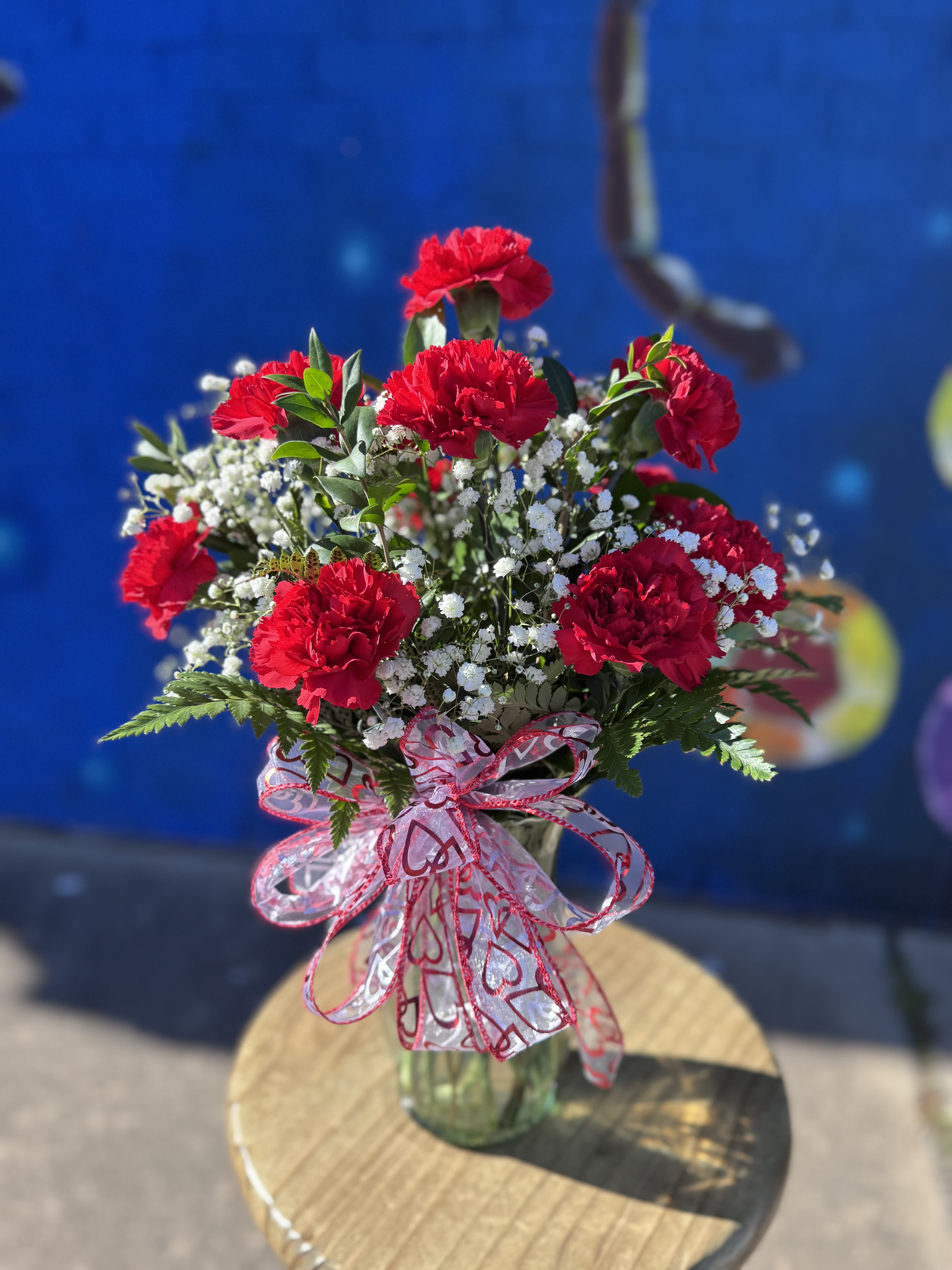 Red carnations in a glass vase with white filler flowers and a heart ribbon