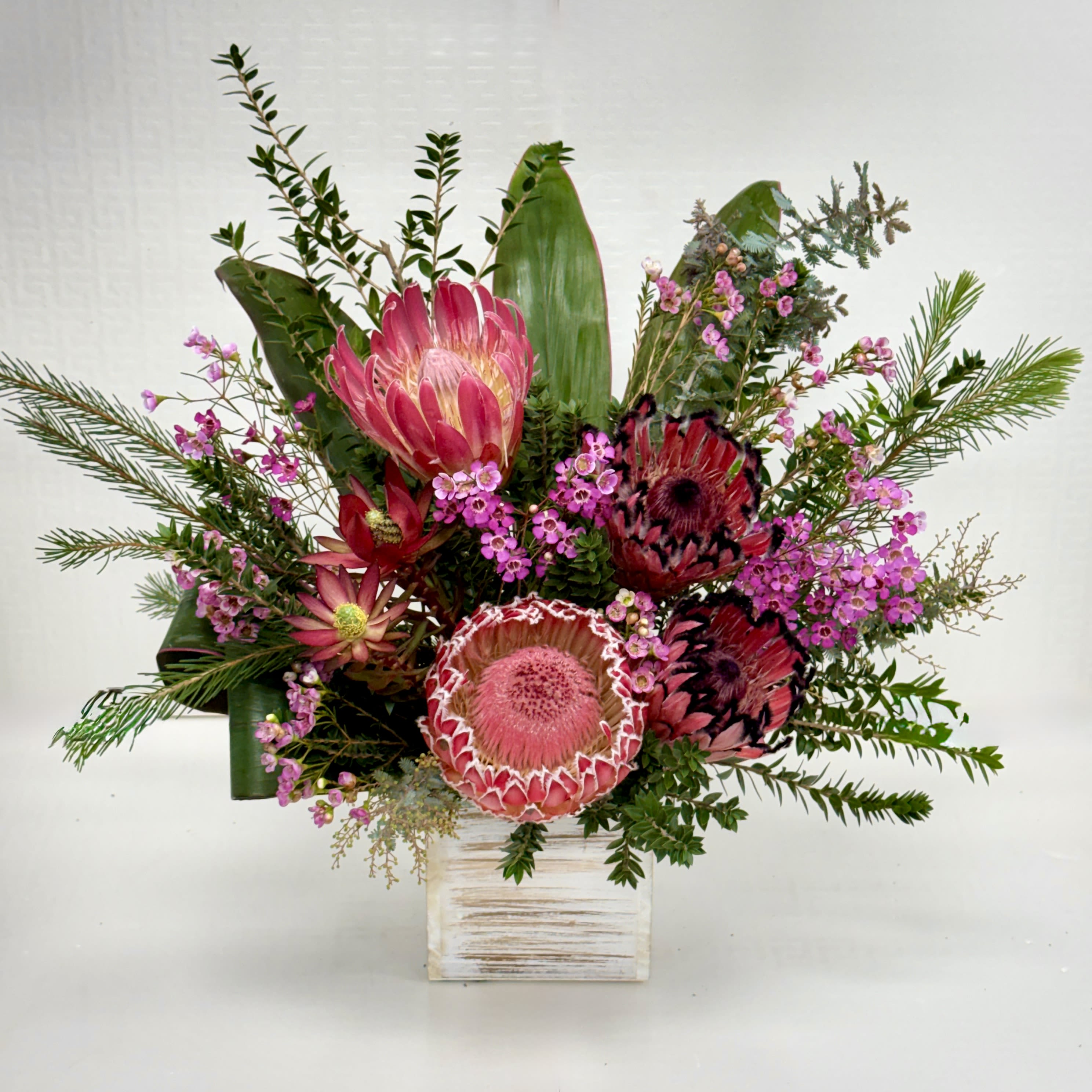 Pink protea and small magenta blooms arranged in a rustic white wooden cube container