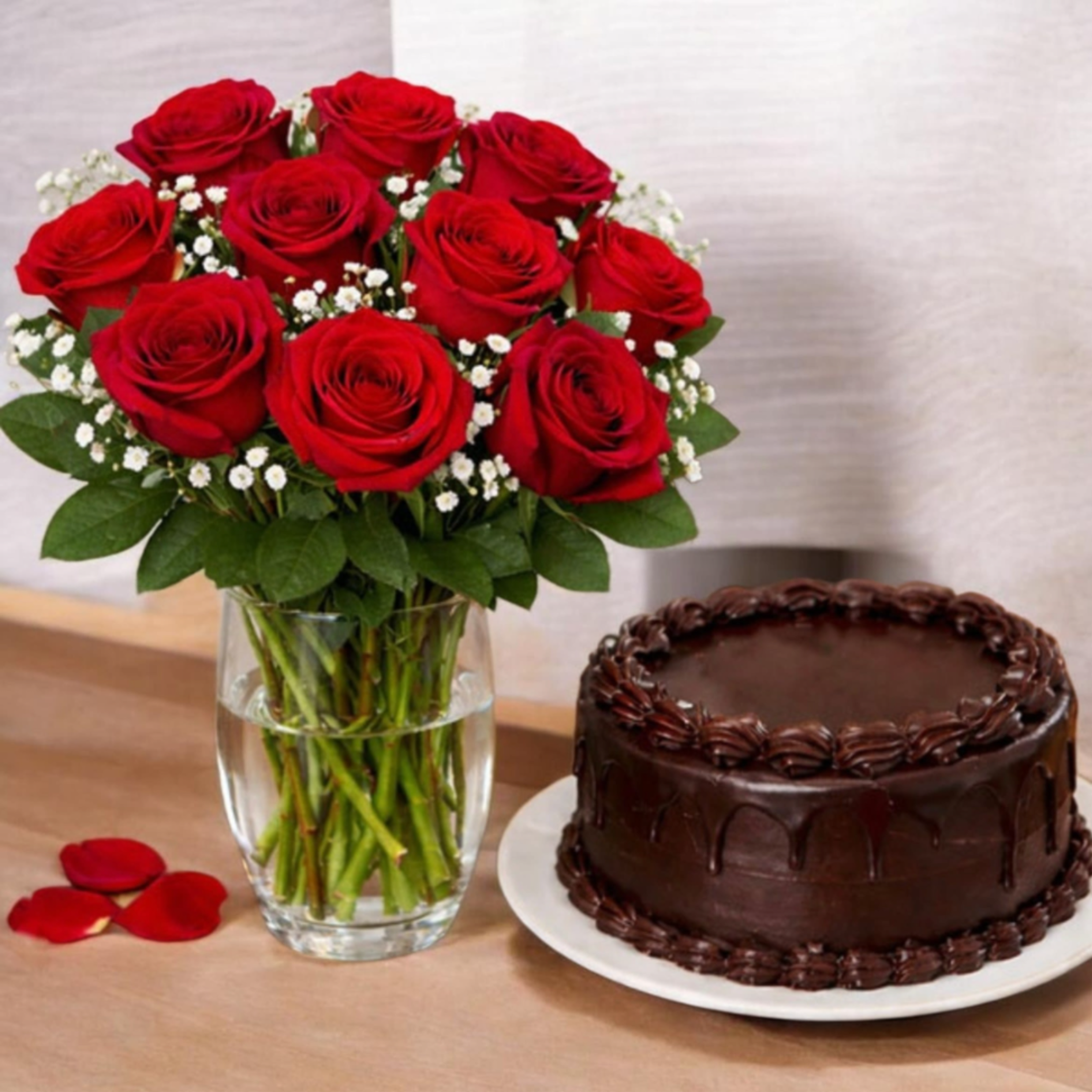 Red Roses Combo Choco - A lush cluster of deep red roses takes center stage in this anniversary bouquet and gift set, the rich petals and rounded shape making an immediate romantic statement. Small white filler flowers and fresh greenery soften the dramatic red, creating a timeless red roses arrangement perfect for Valentine's Day, an anniversary, or any moment you want your person to feel deeply cherished.  Arranged tightly in a clear glass vase, the bouquet presents a full, symmetrical silhouette that reads classic and elegant from every angle. The set includes a chocolate cake served on a white plate beside the vase, with a few scattered rose petals for an intimate, celebratory presentation-ideal for surprising a partner or marking a milestone. Order in time for the occasion and the vase makes a lovely keepsake; refresh stems with water daily and remove spent blooms to help the roses stay beautiful longer.