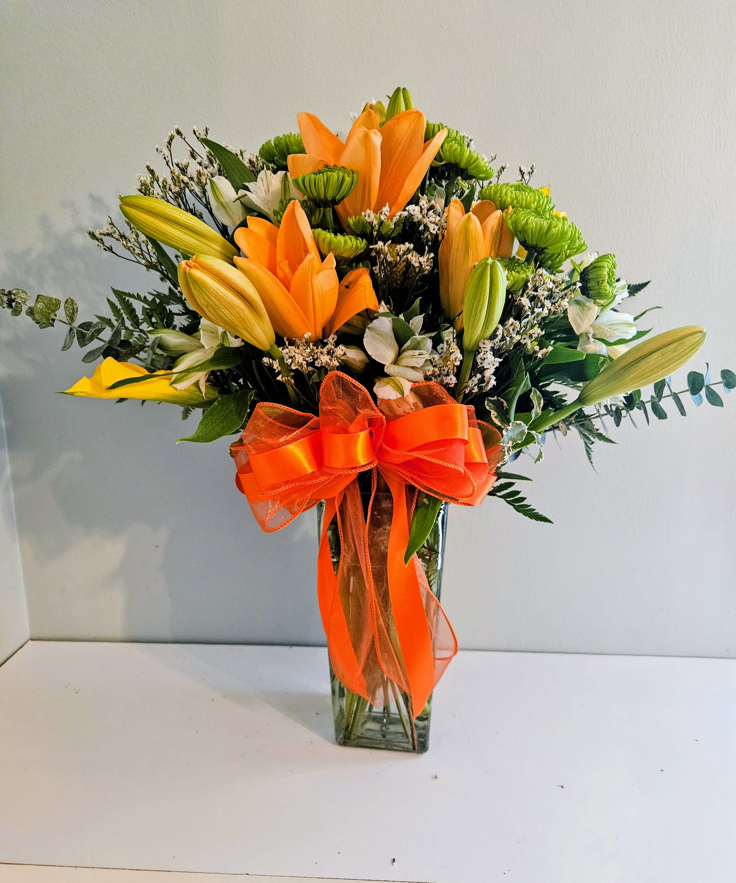 Orange lilies and green chrysanthemums in a clear glass vase with an orange ribbon