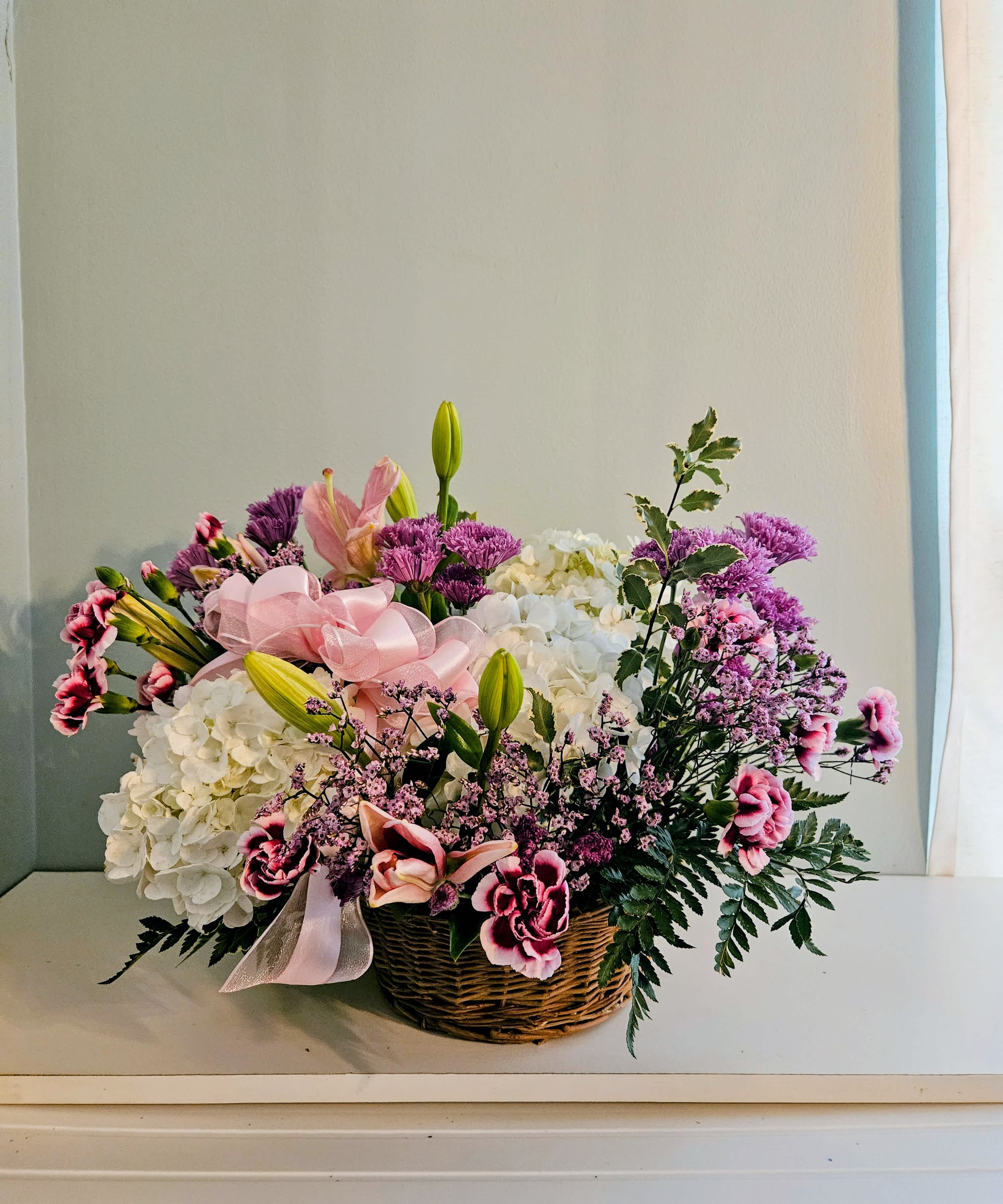 Basket arrangement of white hydrangeas, pink lilies, and purple flowers with a pink ribbon