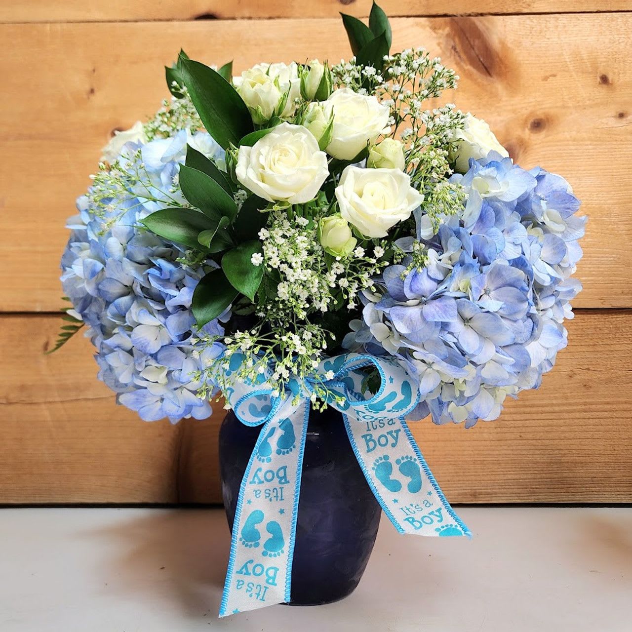 Blue hydrangeas and white roses in a dark vase with a baby boy ribbon